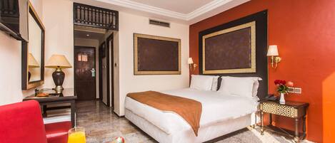 A hotel room with a bed, a desk with a lamp, a chair, a fruit bowl, and a nightstand with a vase.