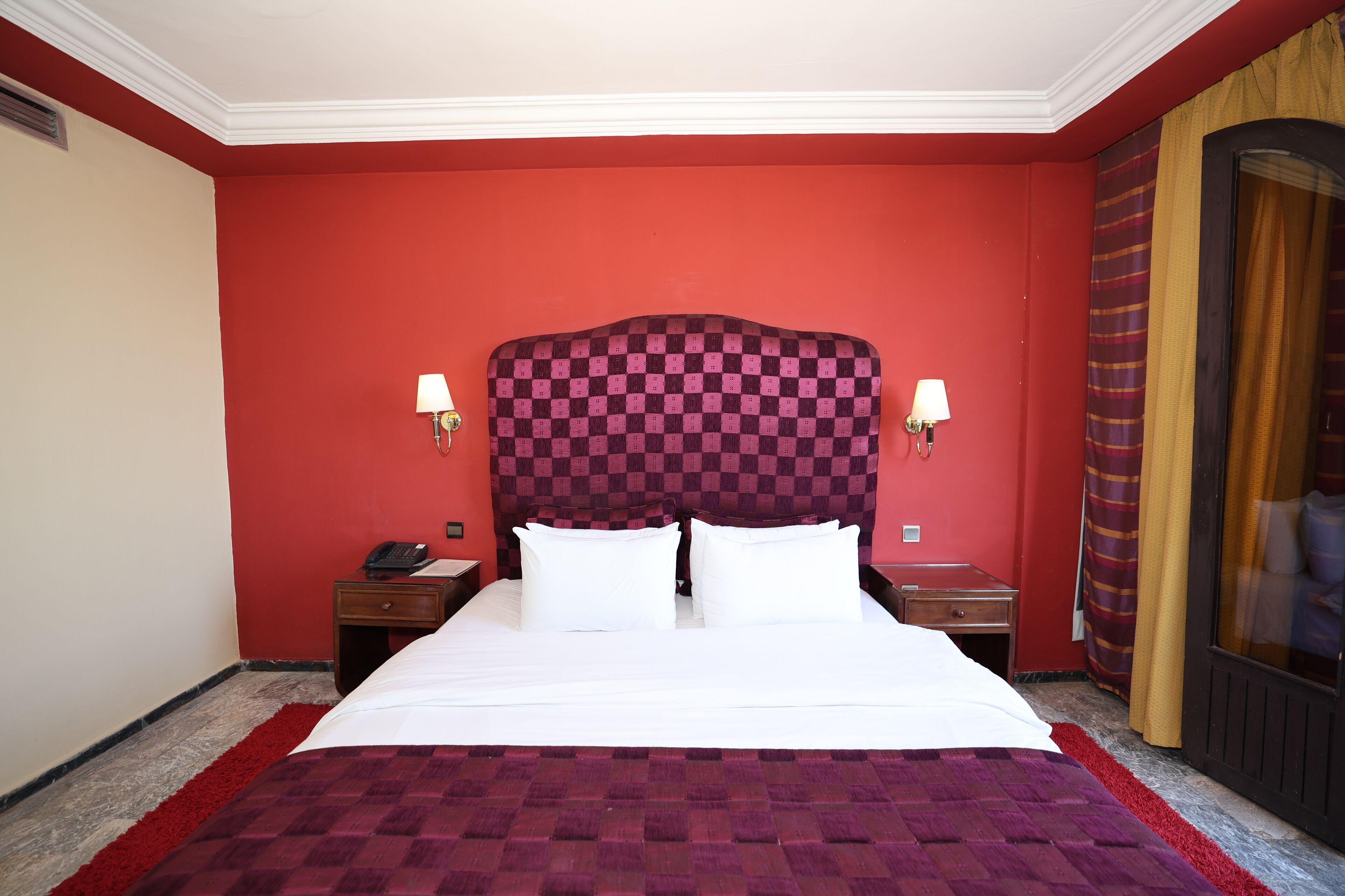 A hotel room with a large bed, red walls, and a checkered headboard. There are two bedside tables with lamps and a telephone on one of them.