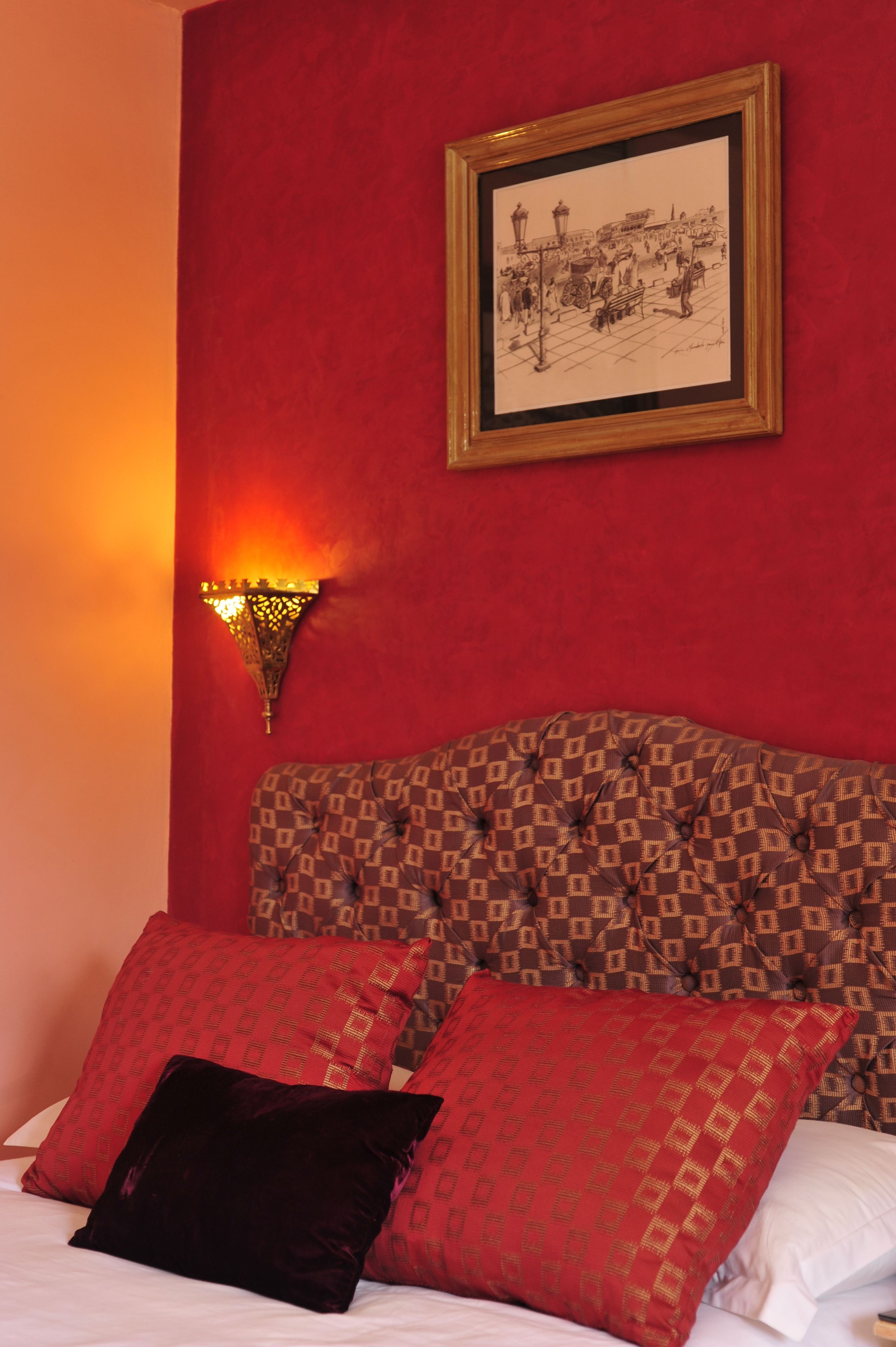 A bedroom with a red wall, a framed picture, a patterned headboard, and a red and black pillow.