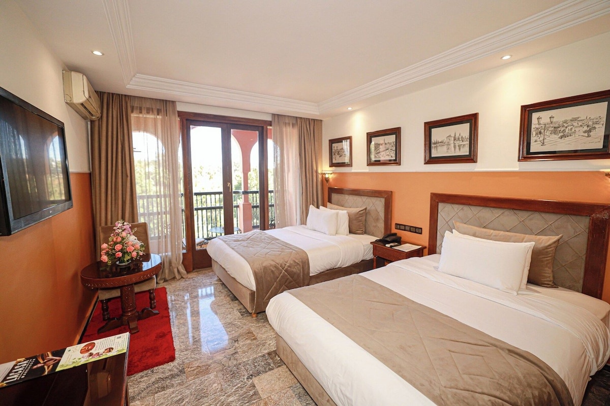 A hotel room with two beds, a desk, a chair, a TV, and a balcony with curtains.