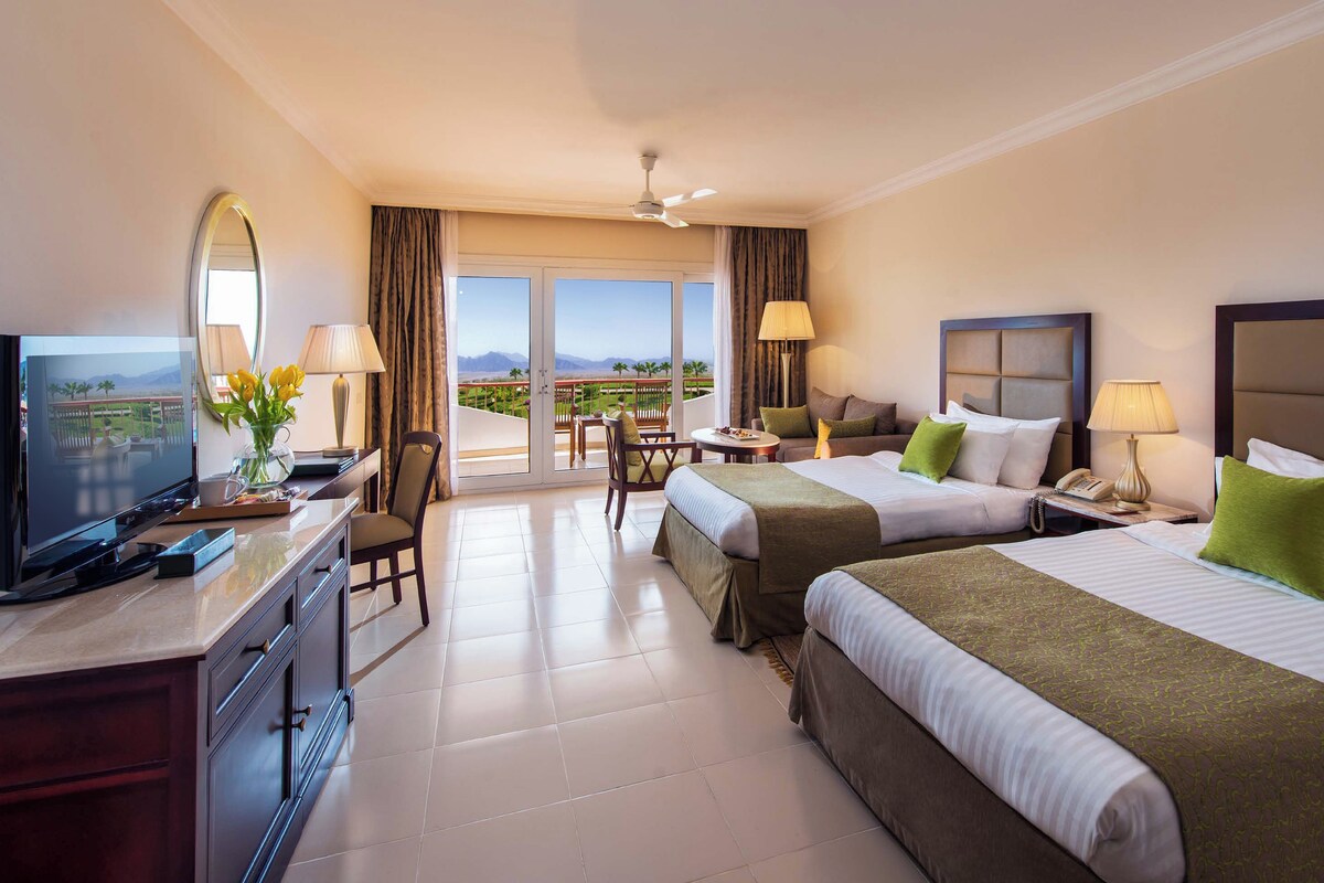 A hotel room with two beds, a desk with a television, a balcony with a view, and a ceiling fan.