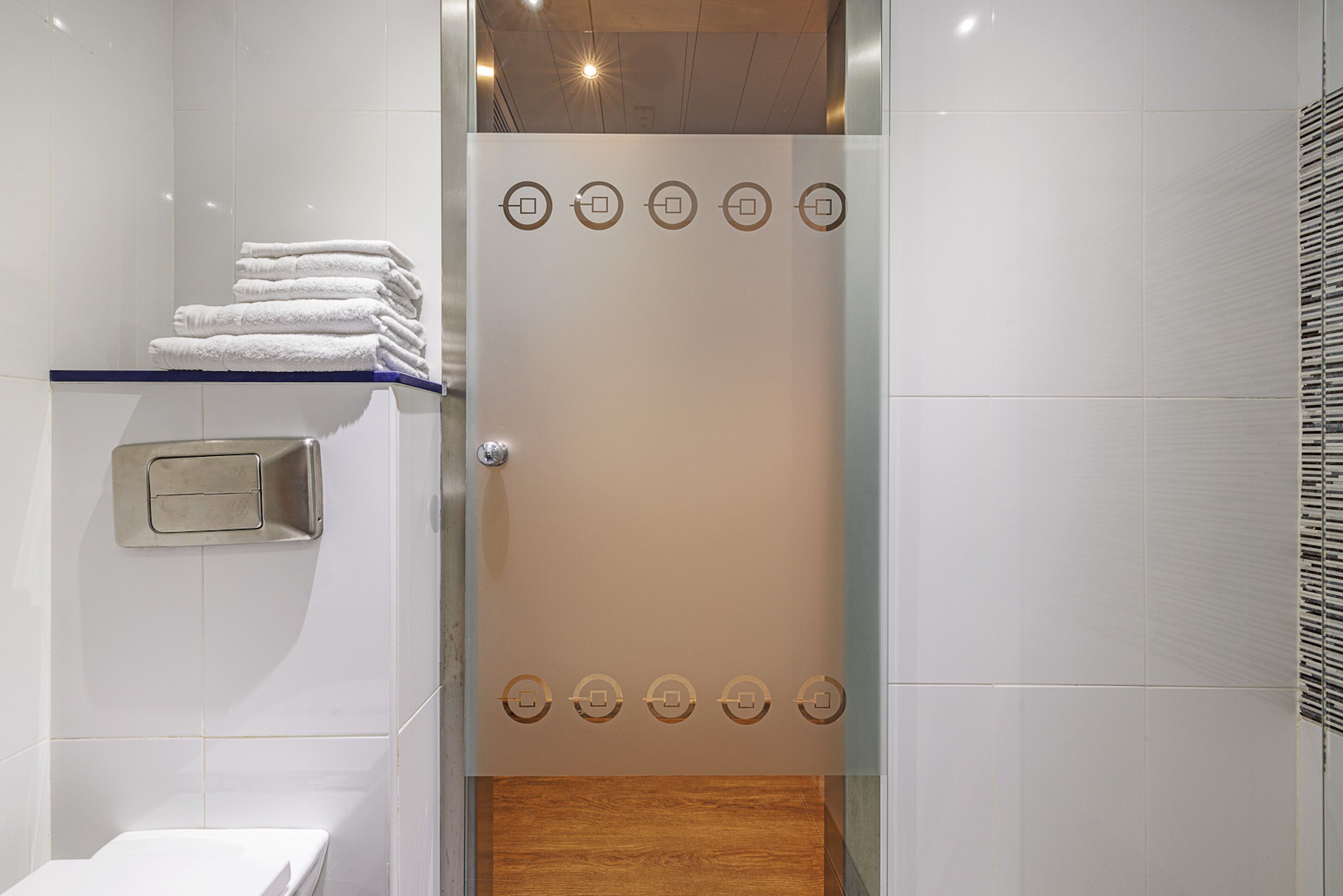 A modern bathroom with a glass shower enclosure, a stack of white towels on a shelf, and a wooden floor.