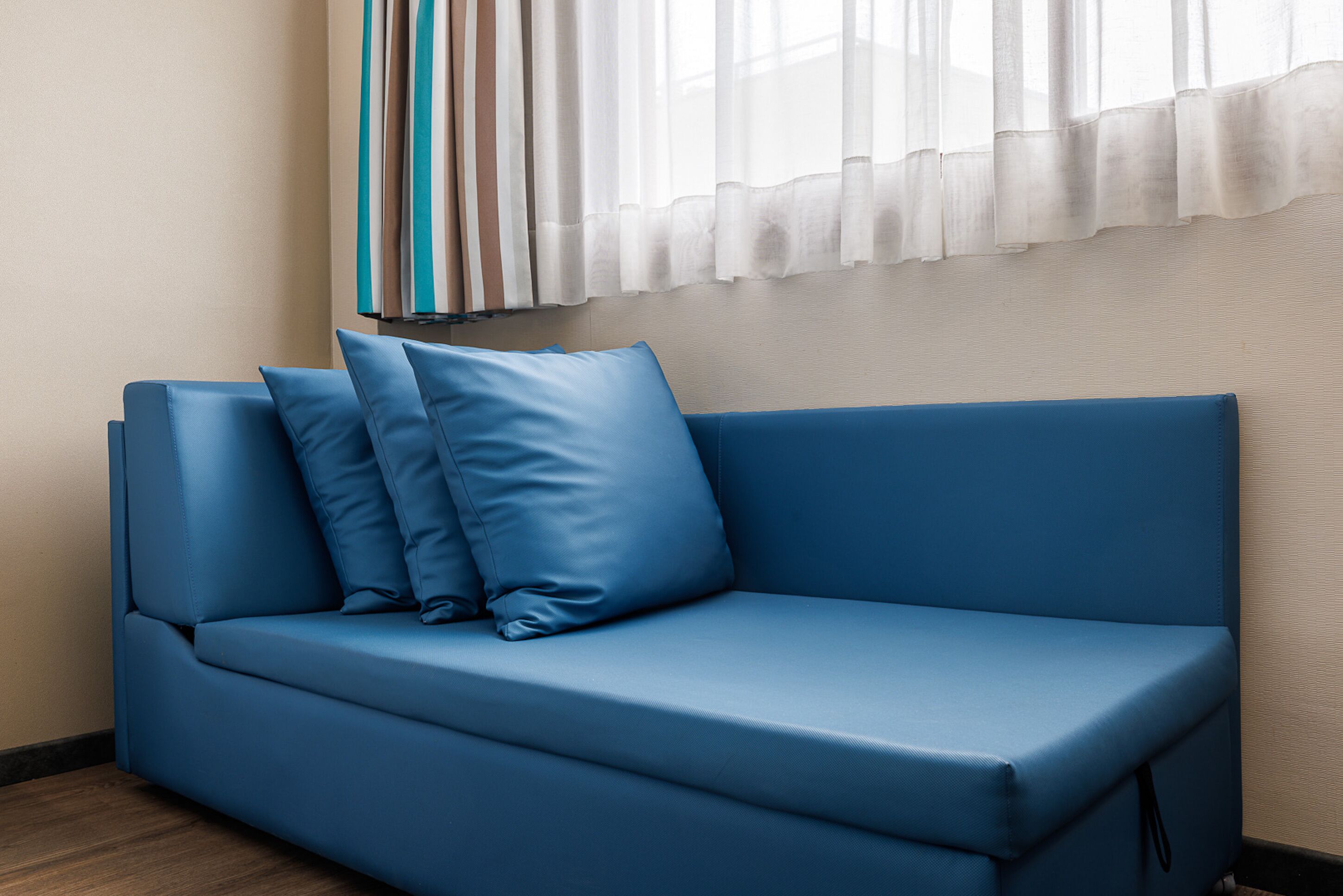 A blue sofa with three cushions in a room with a wooden floor and a window with sheer curtains.