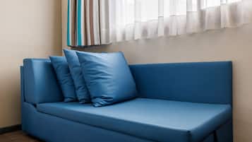 A blue sofa with three cushions in a room with a wooden floor and a window with sheer curtains.