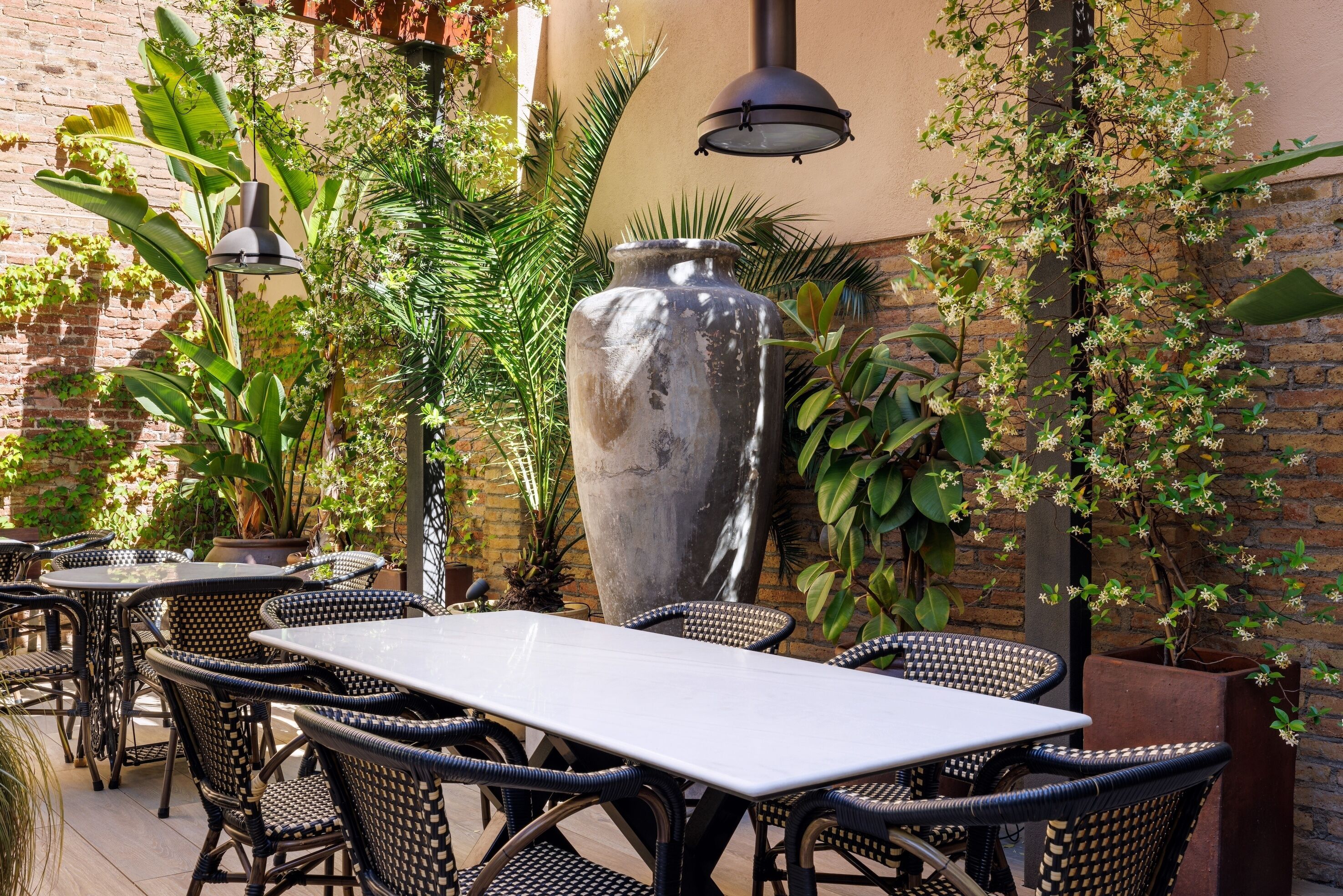 Un patio al aire libre con mesas y sillas de mimbre, rodeado de macetas con plantas y un gran jarrón decorativo.