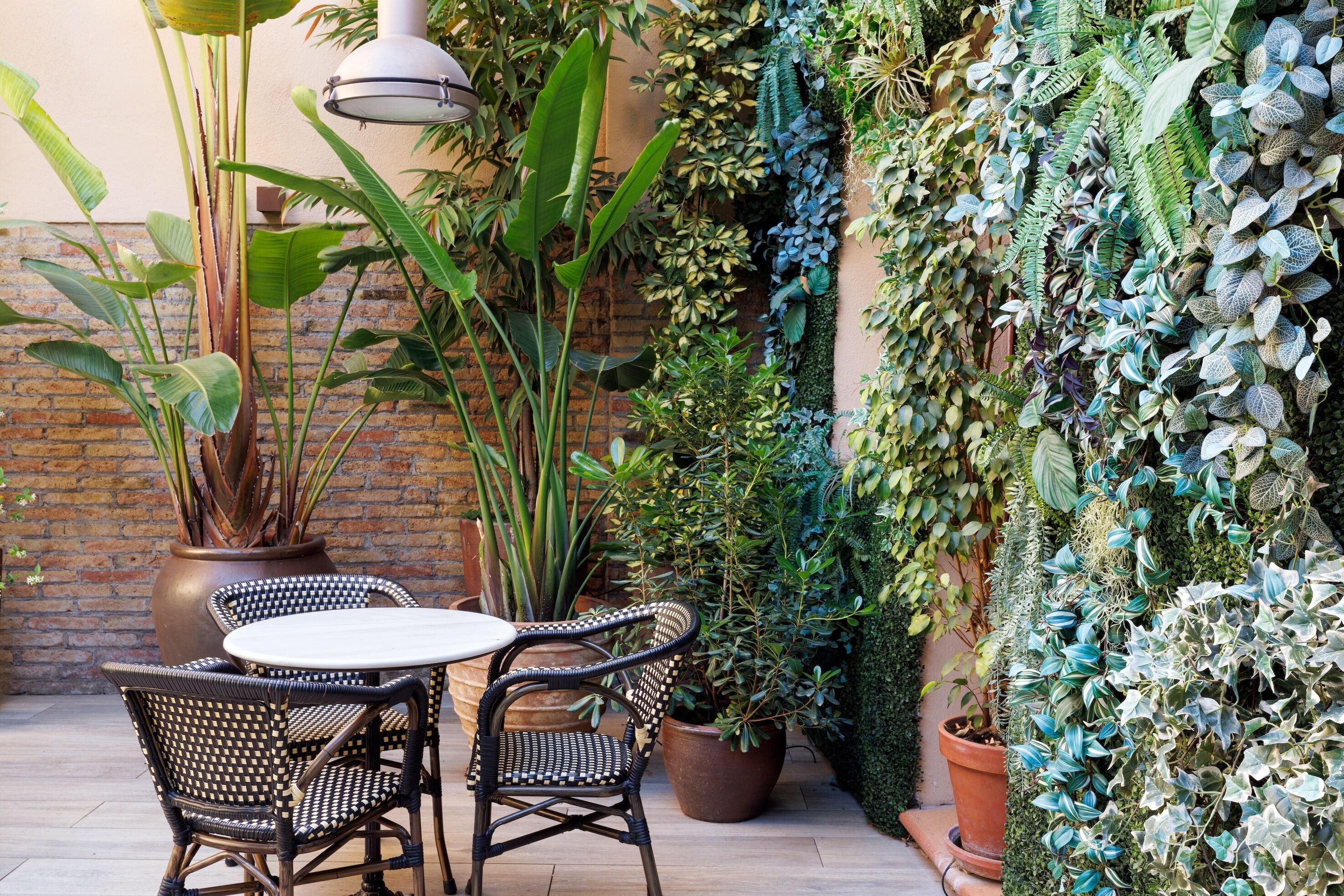 An outdoor patio with wicker chairs and a table, surrounded by various potted plants and a brick wall.