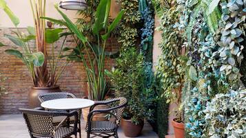 An outdoor patio with wicker chairs and a table, surrounded by various potted plants and a brick wall.