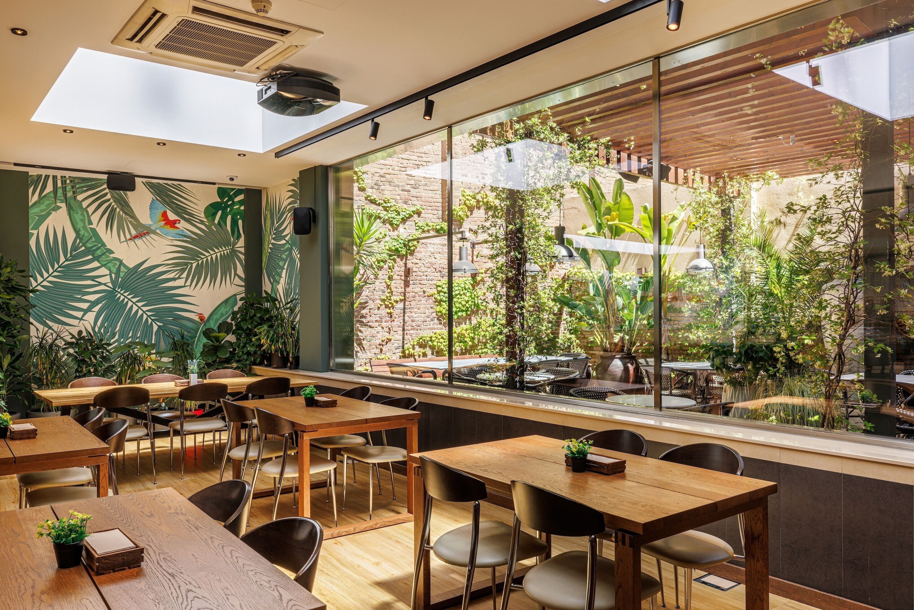 A restaurant with wooden tables and chairs, large windows with a view of an outdoor garden, and a mural of tropical leaves on the wall.