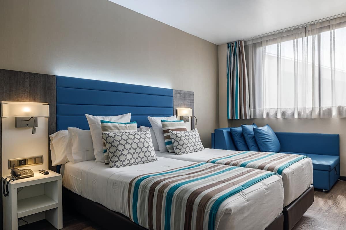 A hotel room with a large bed, two bedside tables, a nightstand, a blue sofa, and a window with sheer curtains.
