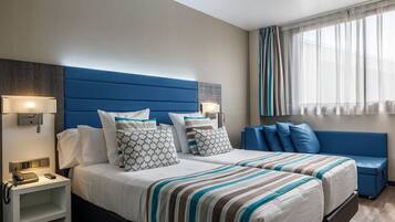 Habitación de hotel con una cama grande, dos mesitas de noche, un buró, un sofá azul y una ventana con cortinas traslúcidas.