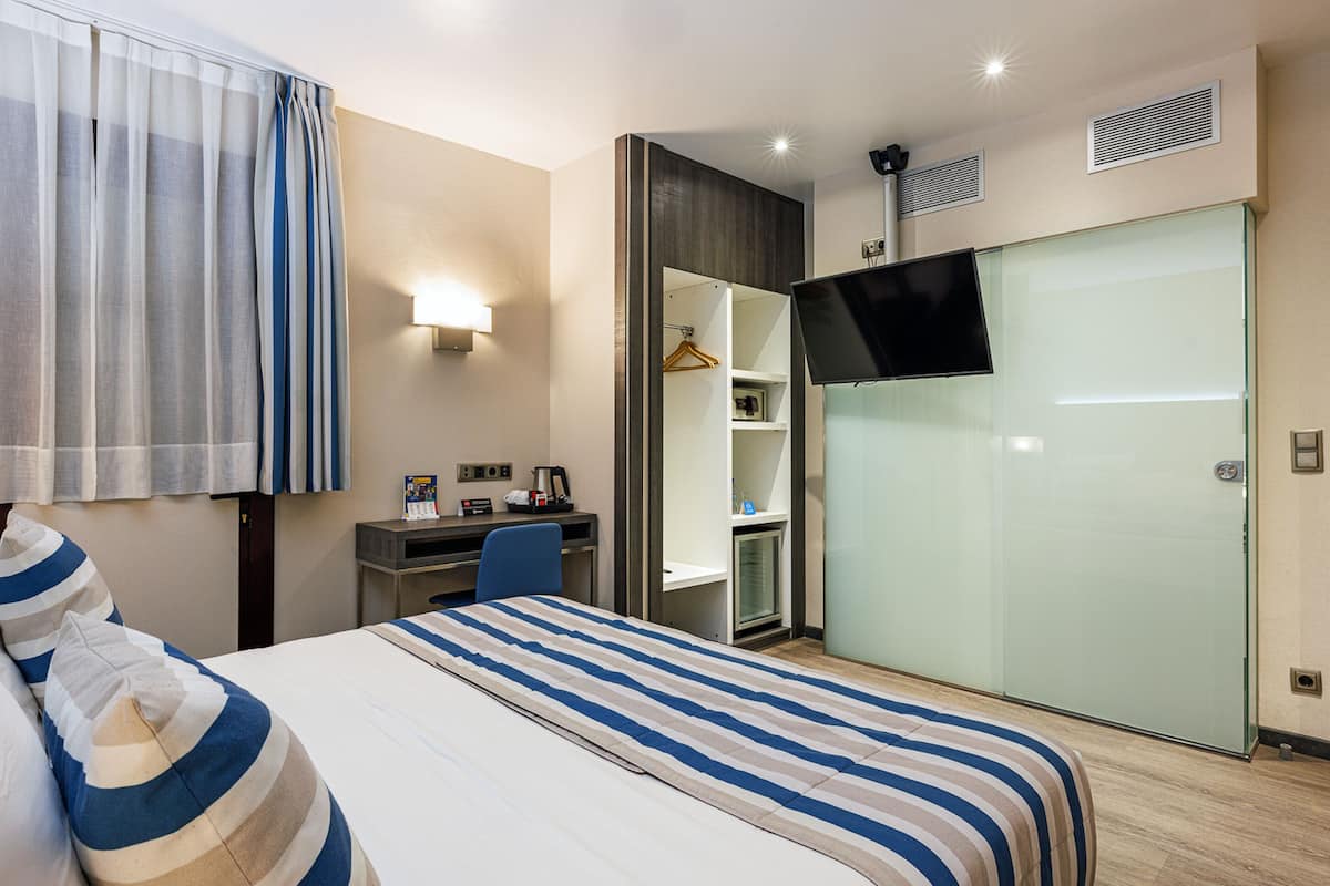 A hotel room with a bed, a desk, a chair, a wardrobe, and a TV.