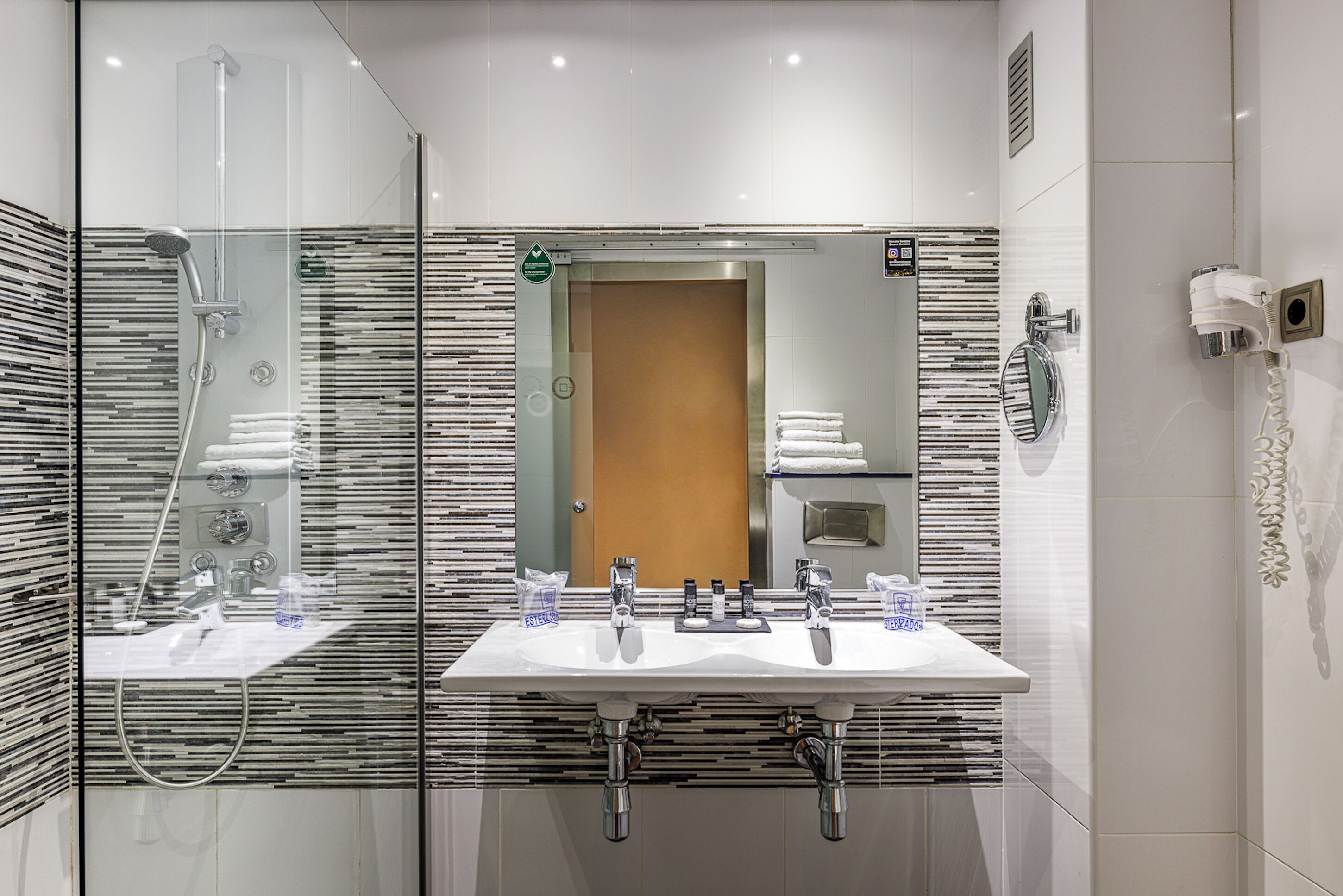 Un baño moderno con ducha de mampara de vidrio, dos lavamanos y un espejo.