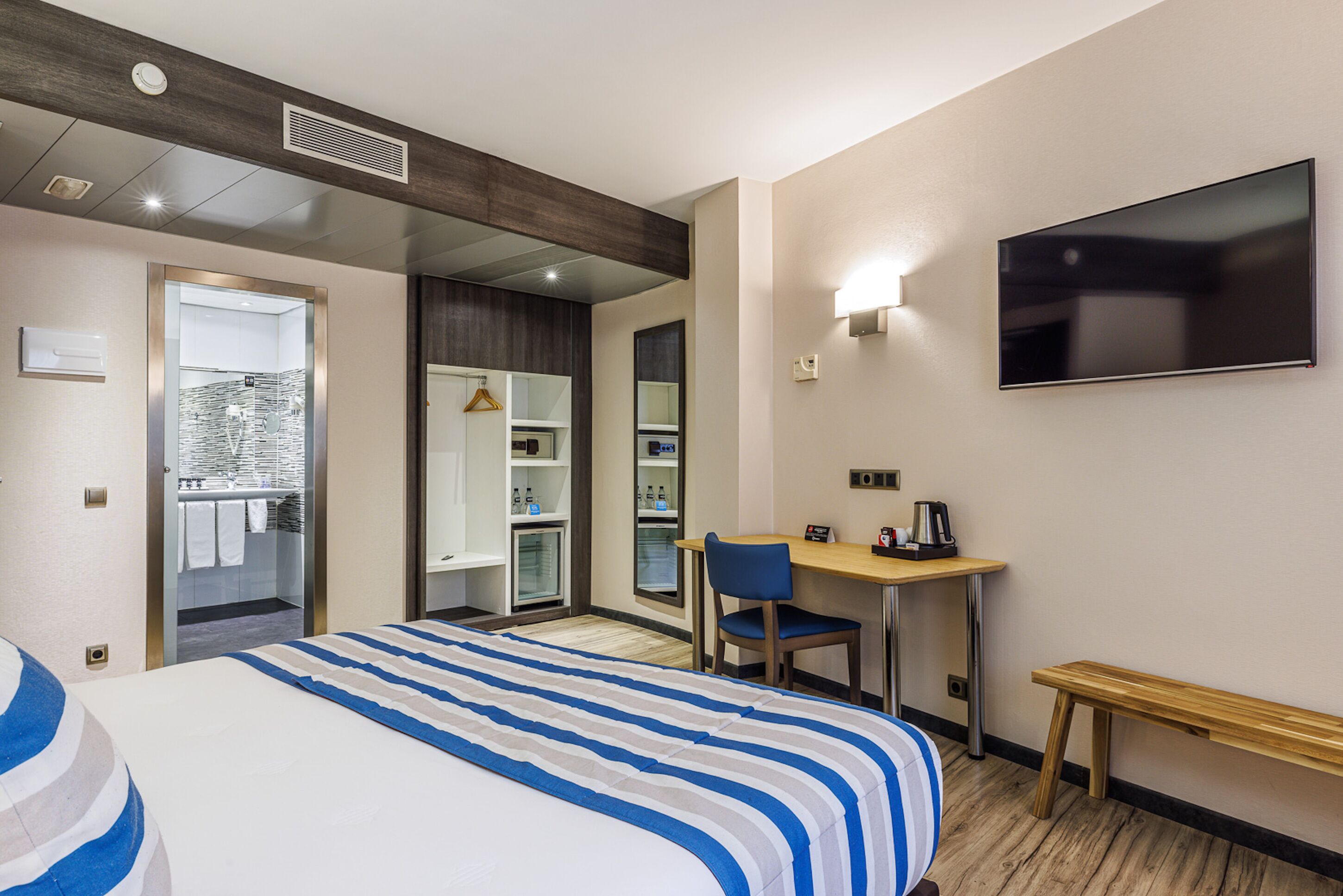 A hotel room with a bed, a desk, a chair, a TV, and a bench.