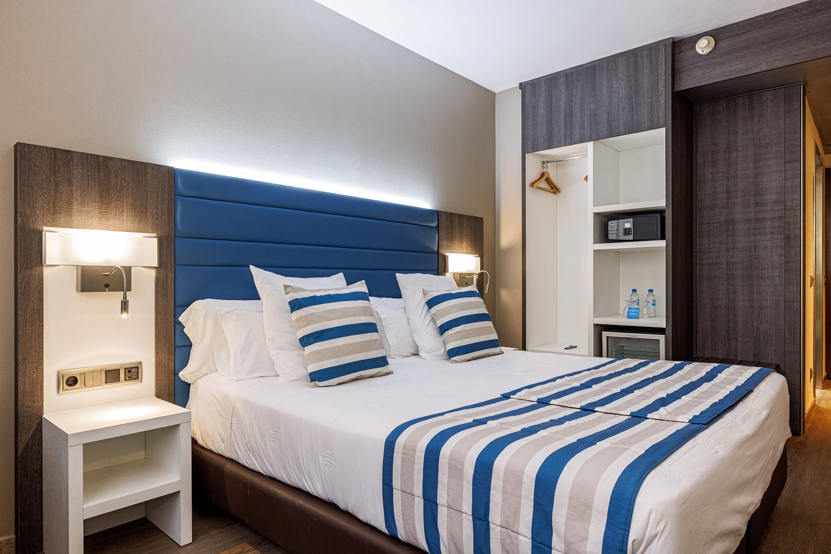 Una habitación de hotel moderna con una cama grande, mesitas de noche con lámparas, un armario empotrado y un escritorio pequeño con un microondas.