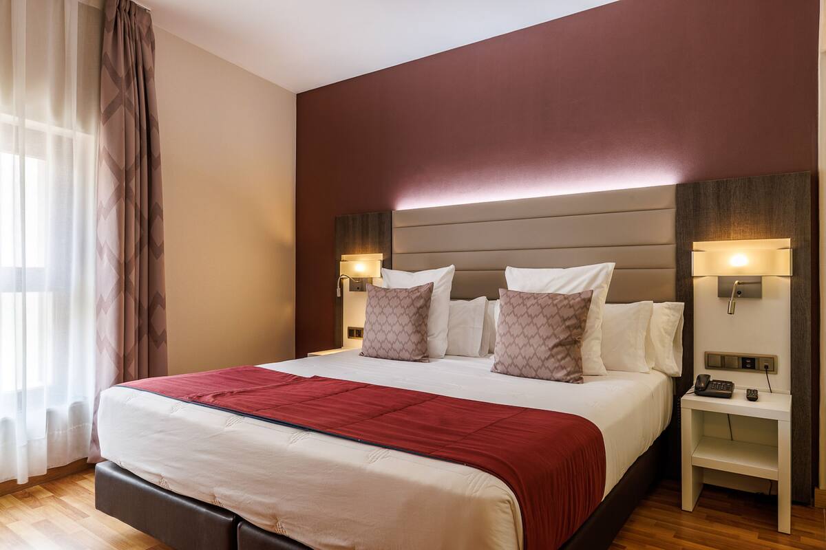 A hotel room with a large bed, two bedside lamps, a nightstand, and a window with curtains.