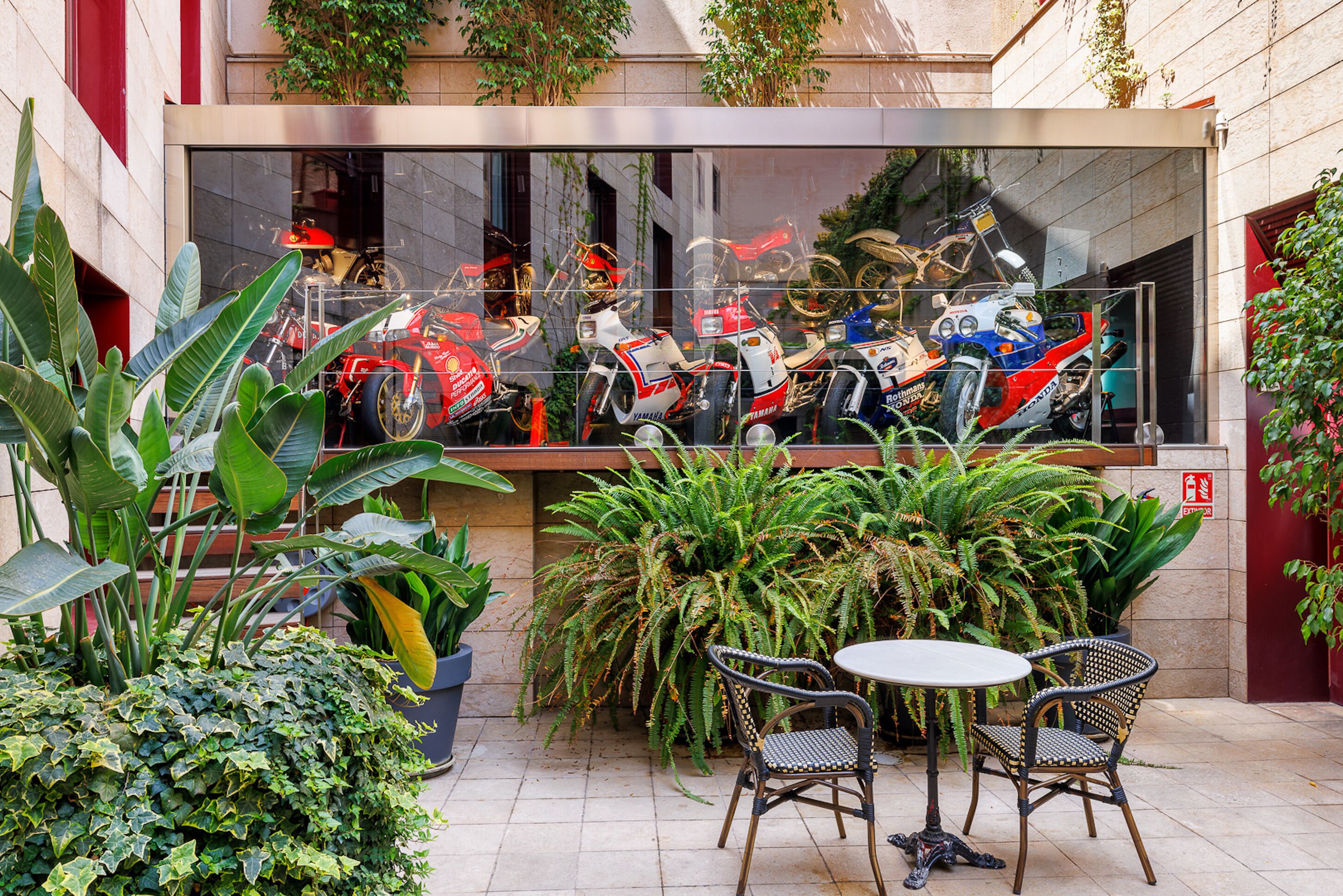 Un patio de restaurante con macetas, dos sillas de mimbre y una vitrina con motocicletas.