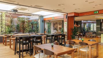 Un amplio comedor interior con varias mesas y sillas de madera, rodeado de grandes ventanales y plantas de interior.