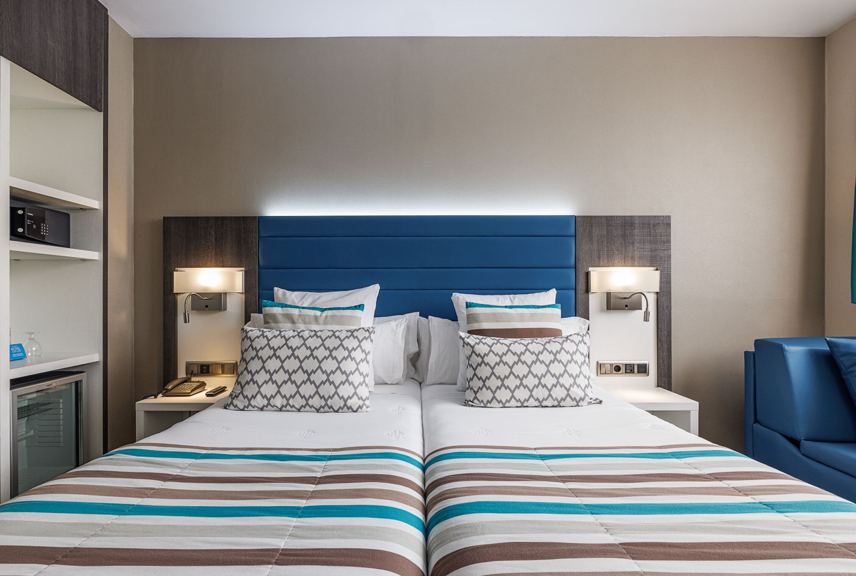 Una cama doble con ropa de cama a rayas, dos mesitas de noche con lámparas, un cabecero azul y un minibar.