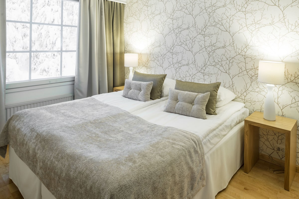 A double bed with a grey blanket, two bedside tables with lamps, a window with curtains, and a wall with a tree branch pattern.
