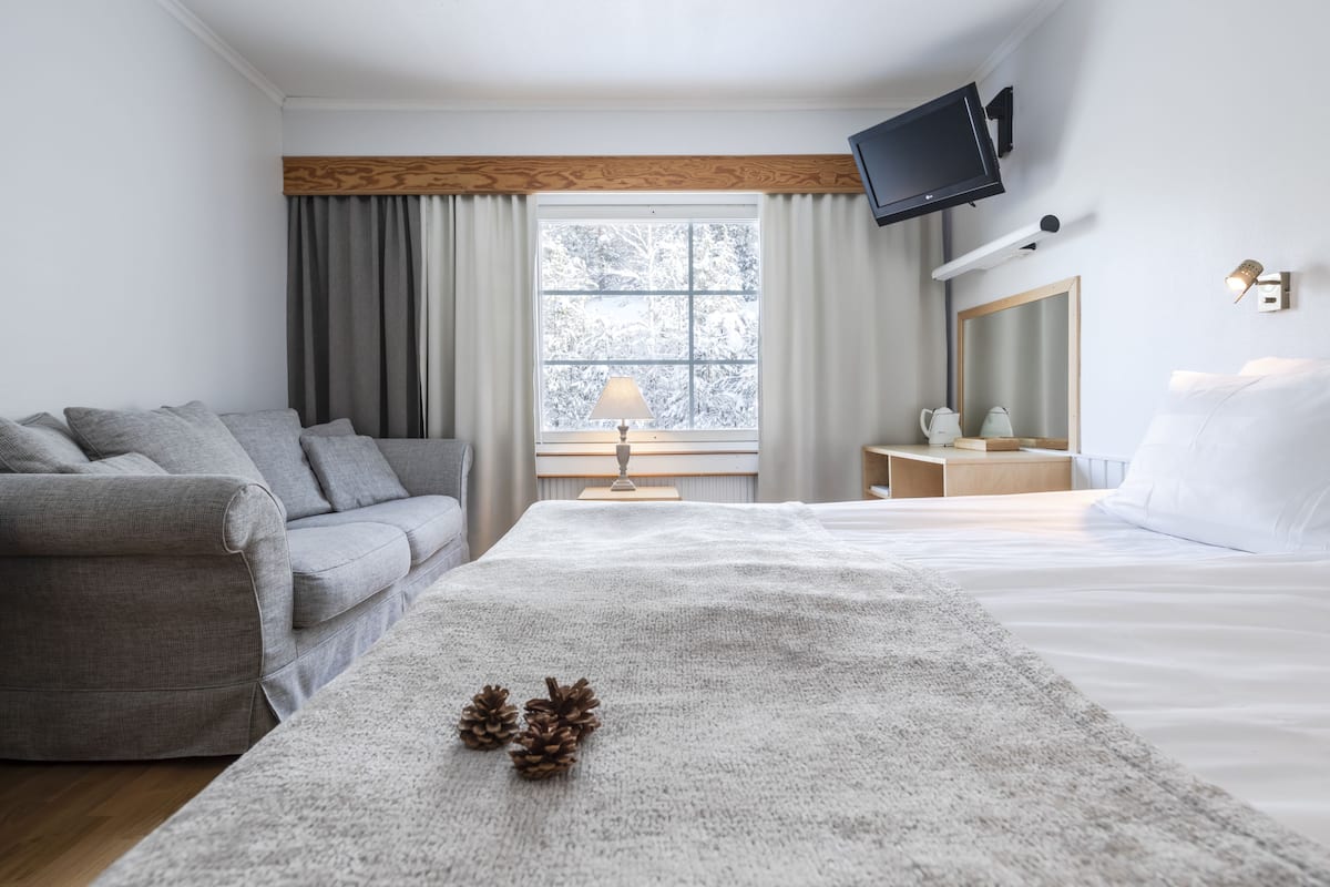 A modern hotel room with a grey sofa, a large bed with white linens, a wooden headboard, a television mounted on the wall, and a window with a view of snow-covered trees.