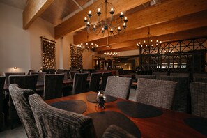 Een restaurant met houten balken, ronde tafels met zwarte placemats en een kroonluchter.