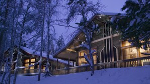 Una cabaña cubierta de nieve con fachada de madera y ventanas amplias, rodeada de árboles.