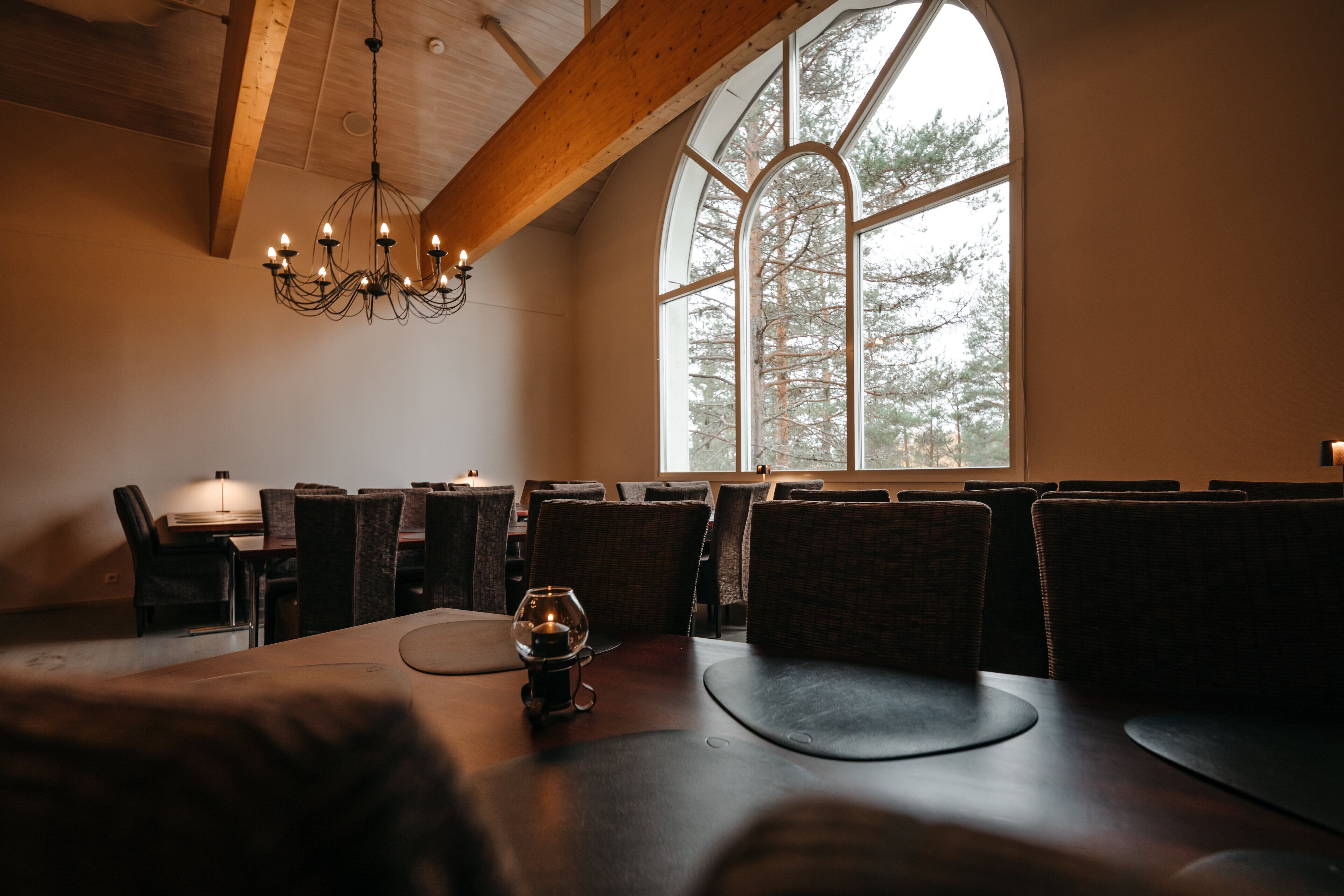 Een restaurant met een hoog houten plafond, grote gewelfde ramen en een kroonluchter. Tafels zijn gedekt met stoelen, en op één tafel staat een wijnglas.