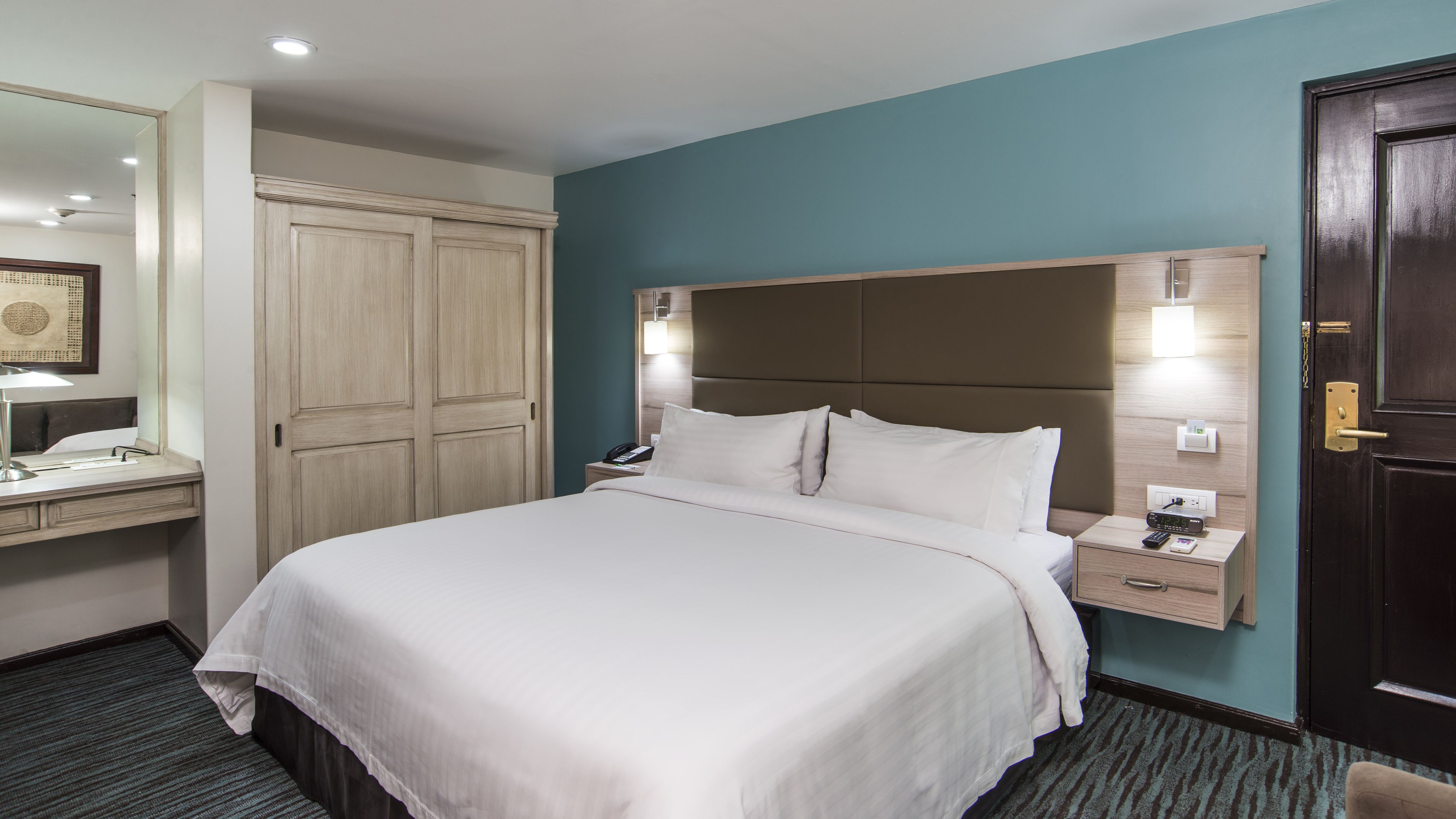 Una habitación de hotel con una cama grande, un escritorio con lámpara, un armario y una mesita de noche con teléfono y control remoto.
