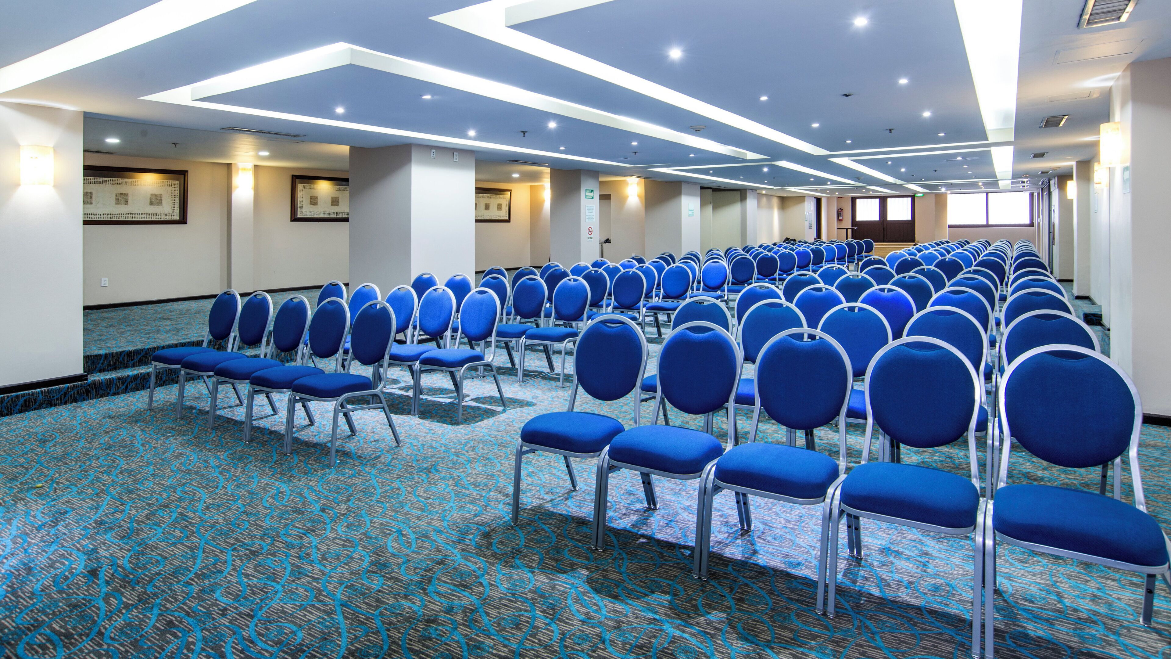 A conference room with rows of blue chairs, a patterned carpet, and a ceiling with recessed lighting.