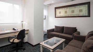 Et hotelværelse med skrivebord, kontorstol, to sofaer, et sofabord med glasplade og en dekorativ vægdekoration.