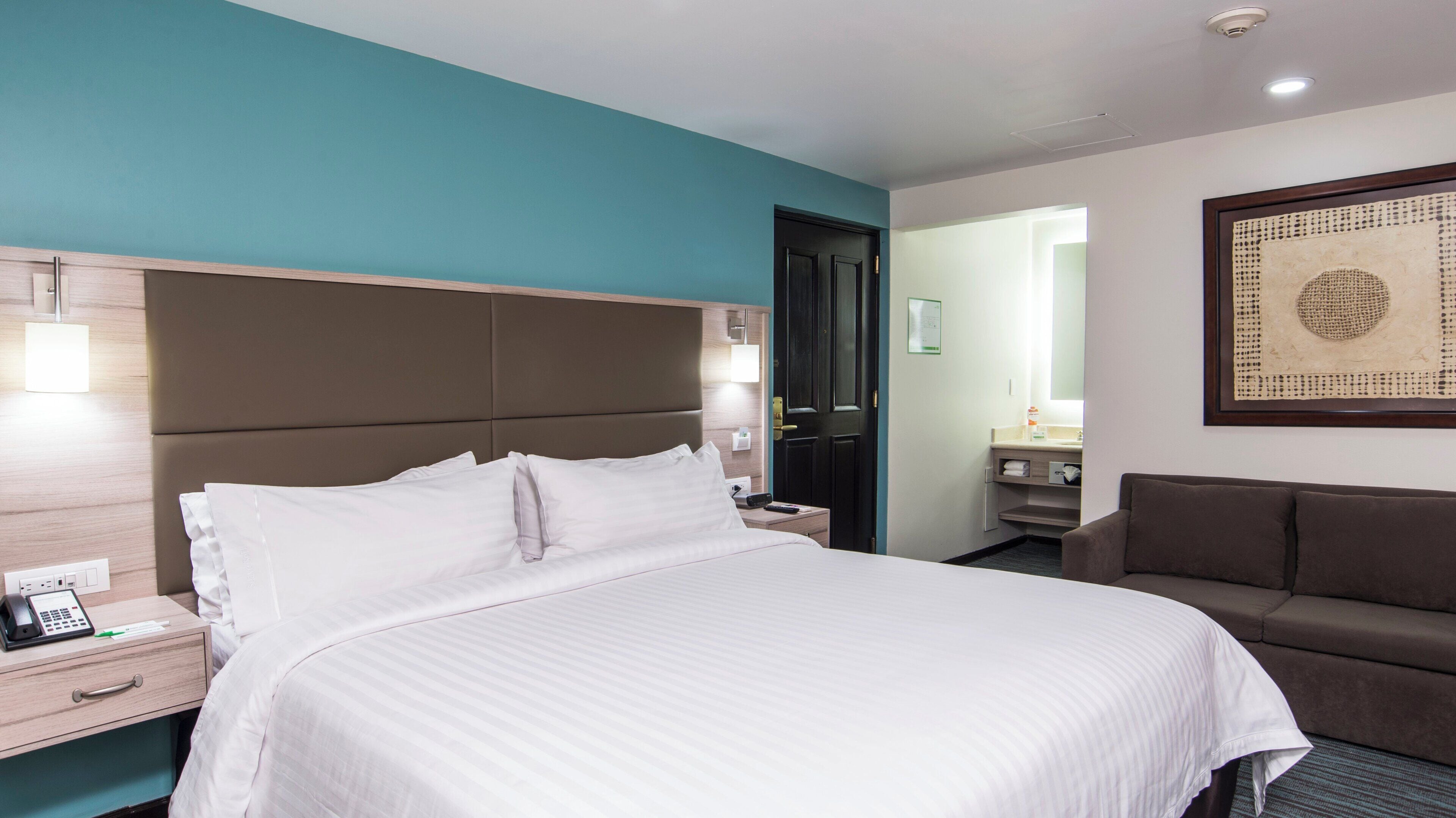 A hotel room with a large bed, a bedside table with a phone, a brown sofa, and a bathroom visible through an open door.