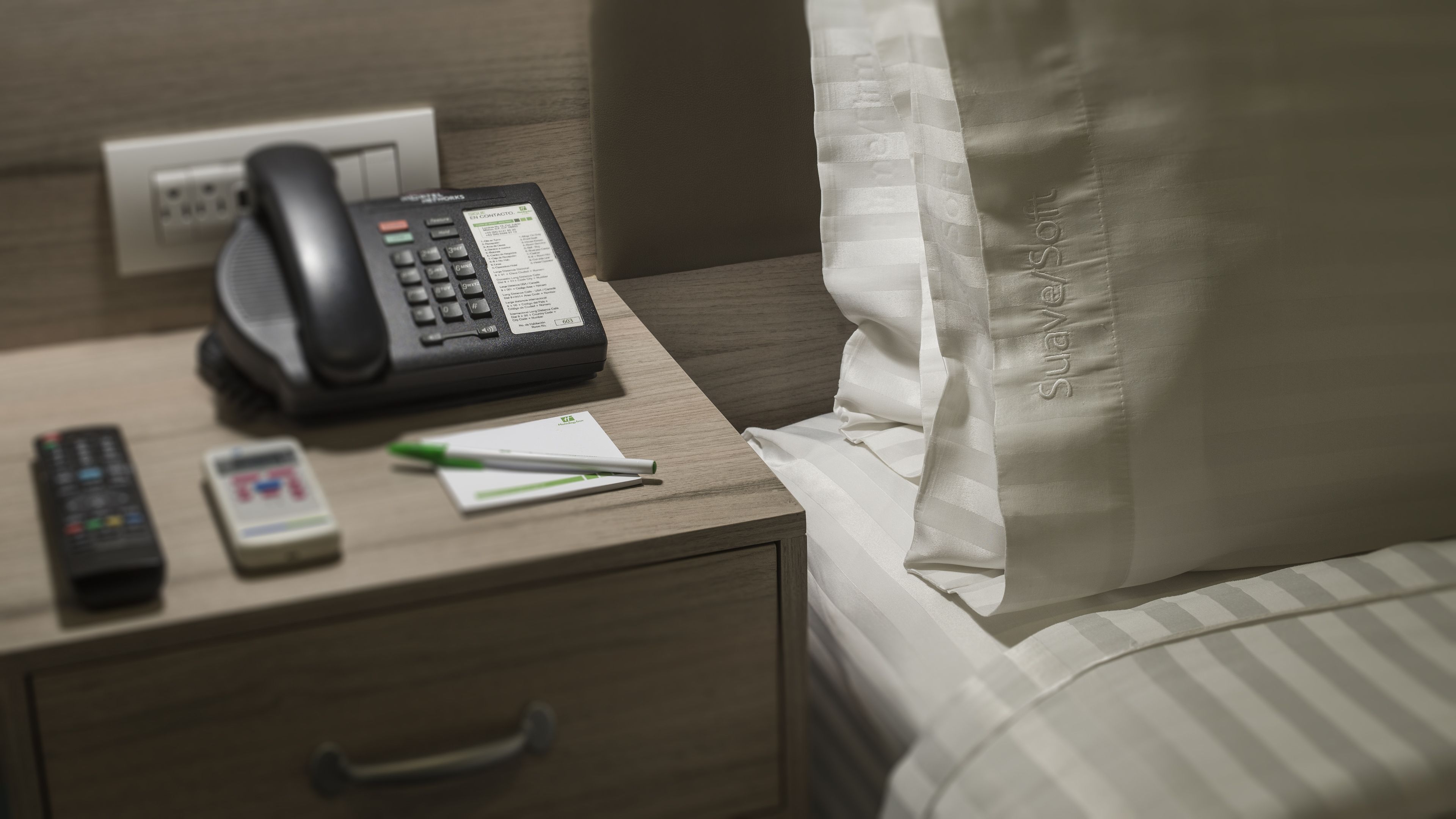 A hotel room with a bedside telephone, a remote control, a pen, and a notepad on the nightstand.