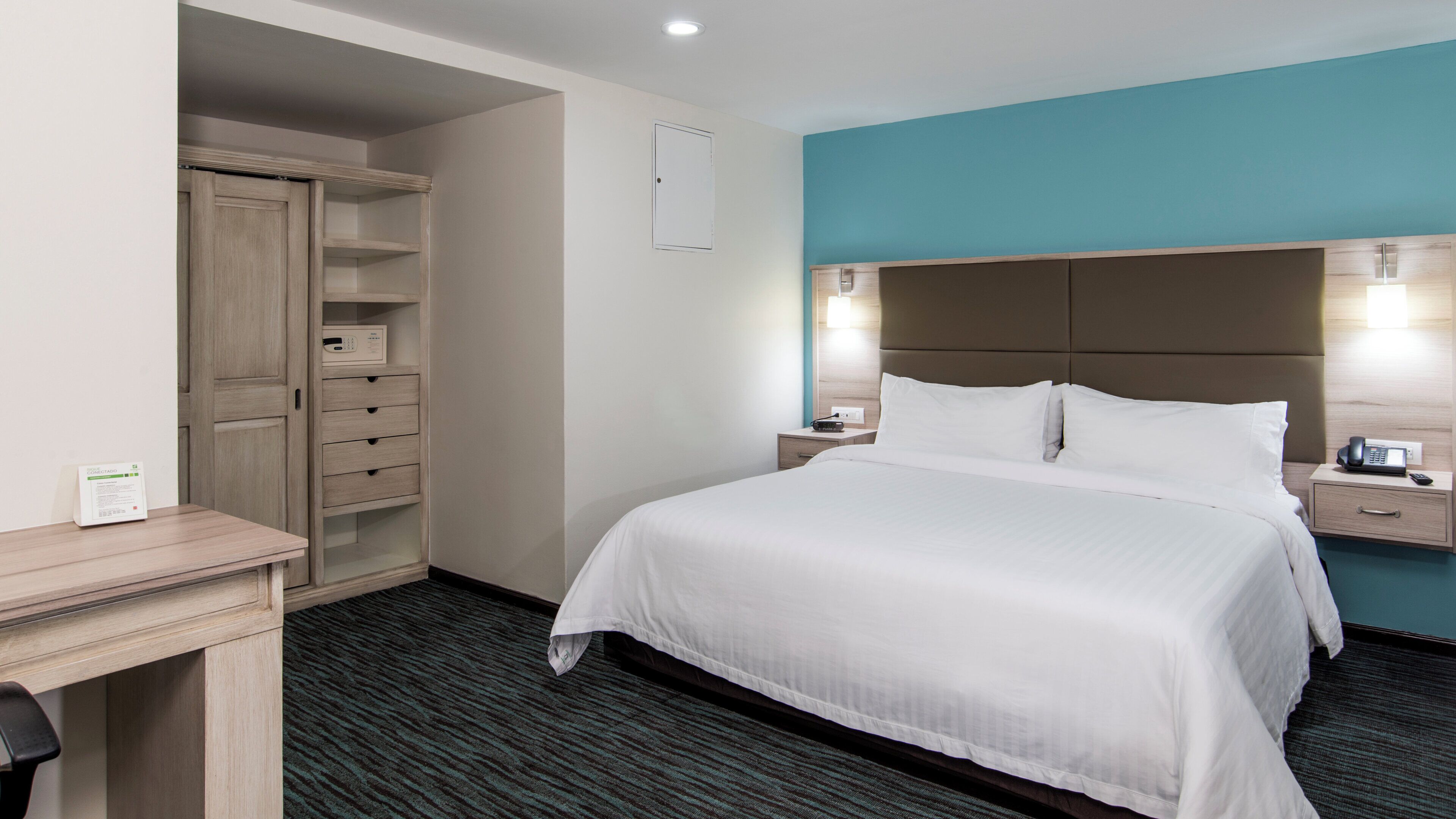 A hotel room with a bed, bedside tables, a desk, a wardrobe, and a wall-mounted light fixture.