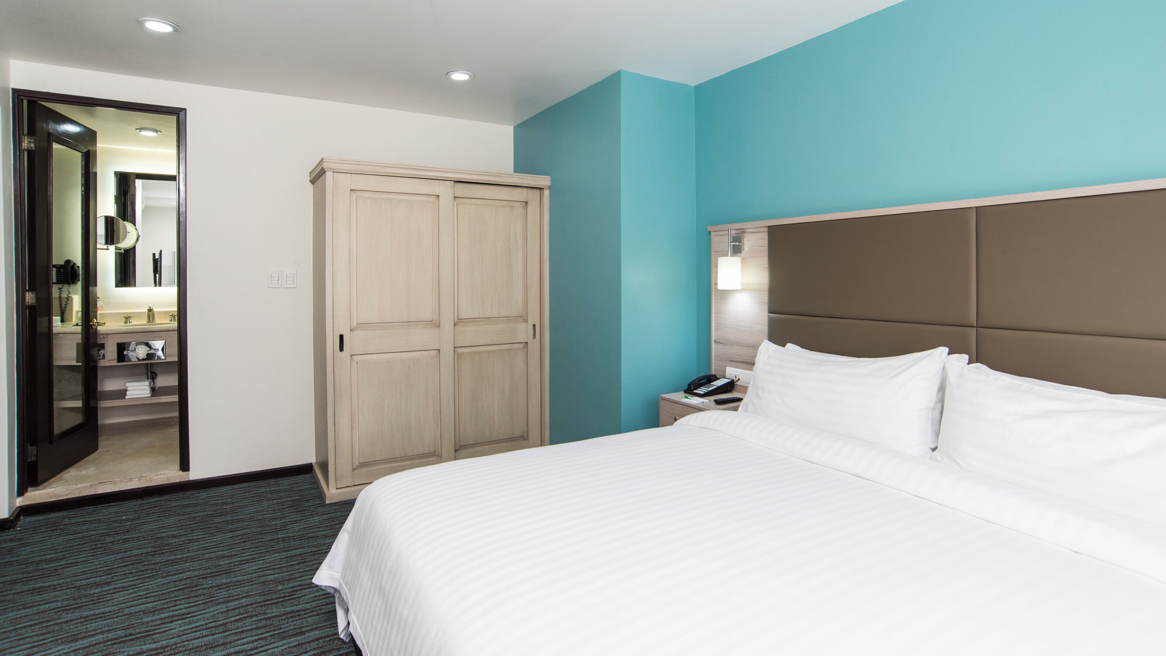 A hotel room with a bed, a wardrobe, a bathroom with a mirror, and a nightstand with a lamp.