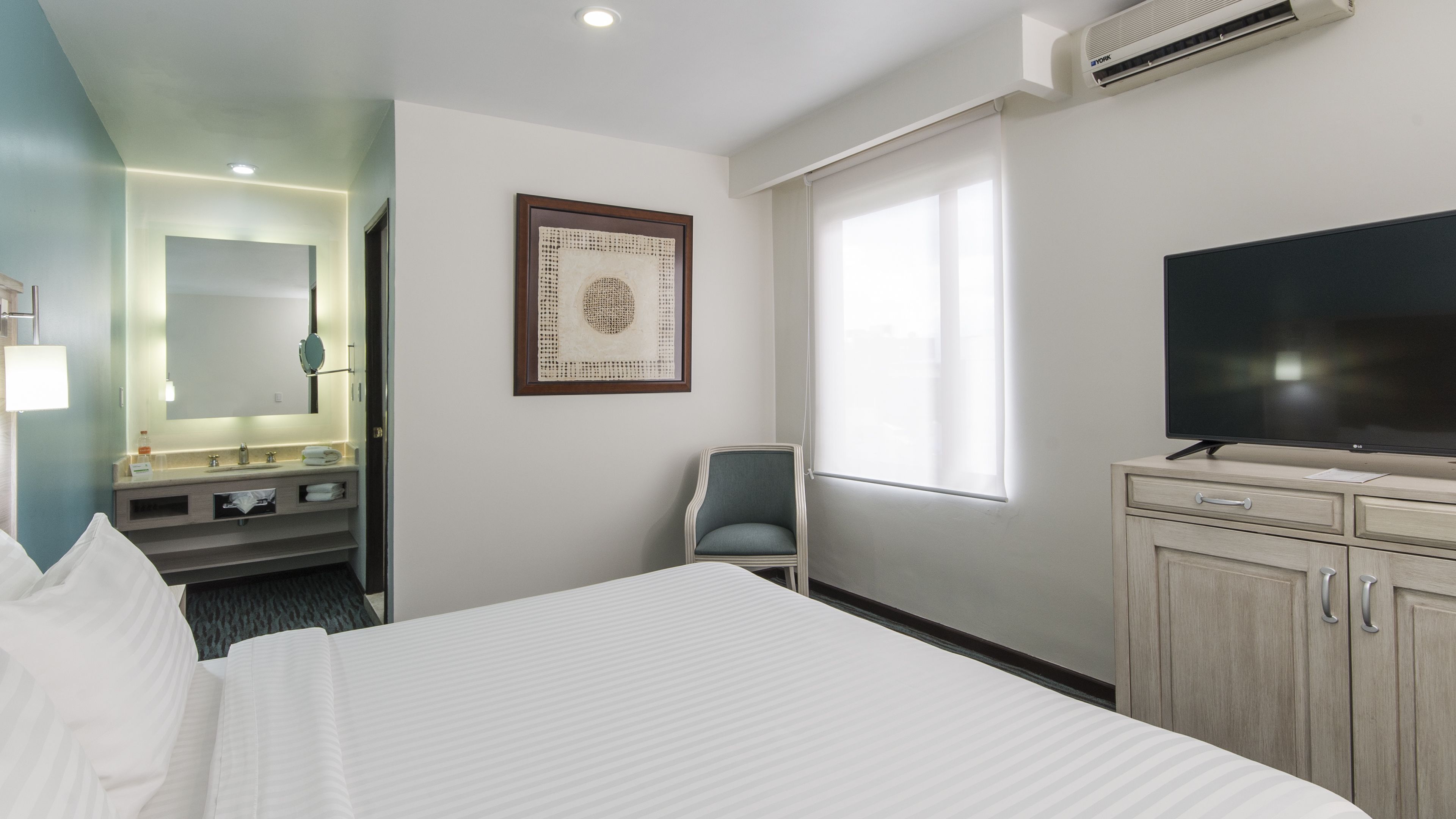 Une chambre d’hôtel avec un grand lit, une télévision, un bureau, une chaise, une salle de bain avec un miroir et une fenêtre.