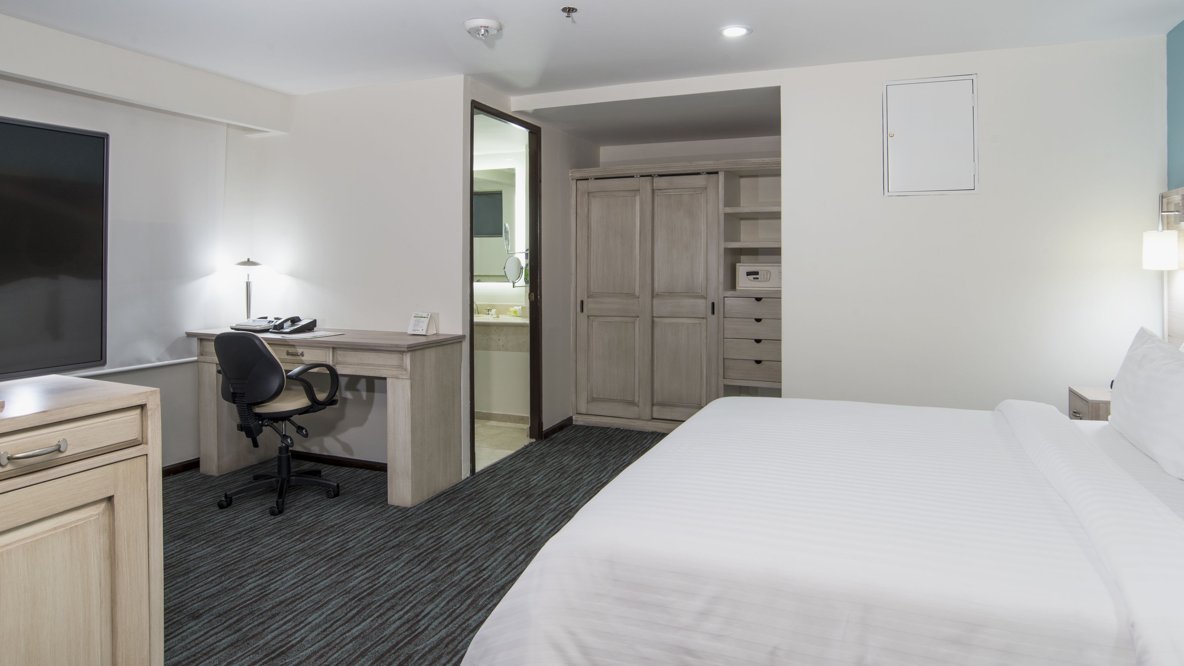A hotel room with a bed, desk, chair, wardrobe, and a bathroom visible through an open door.