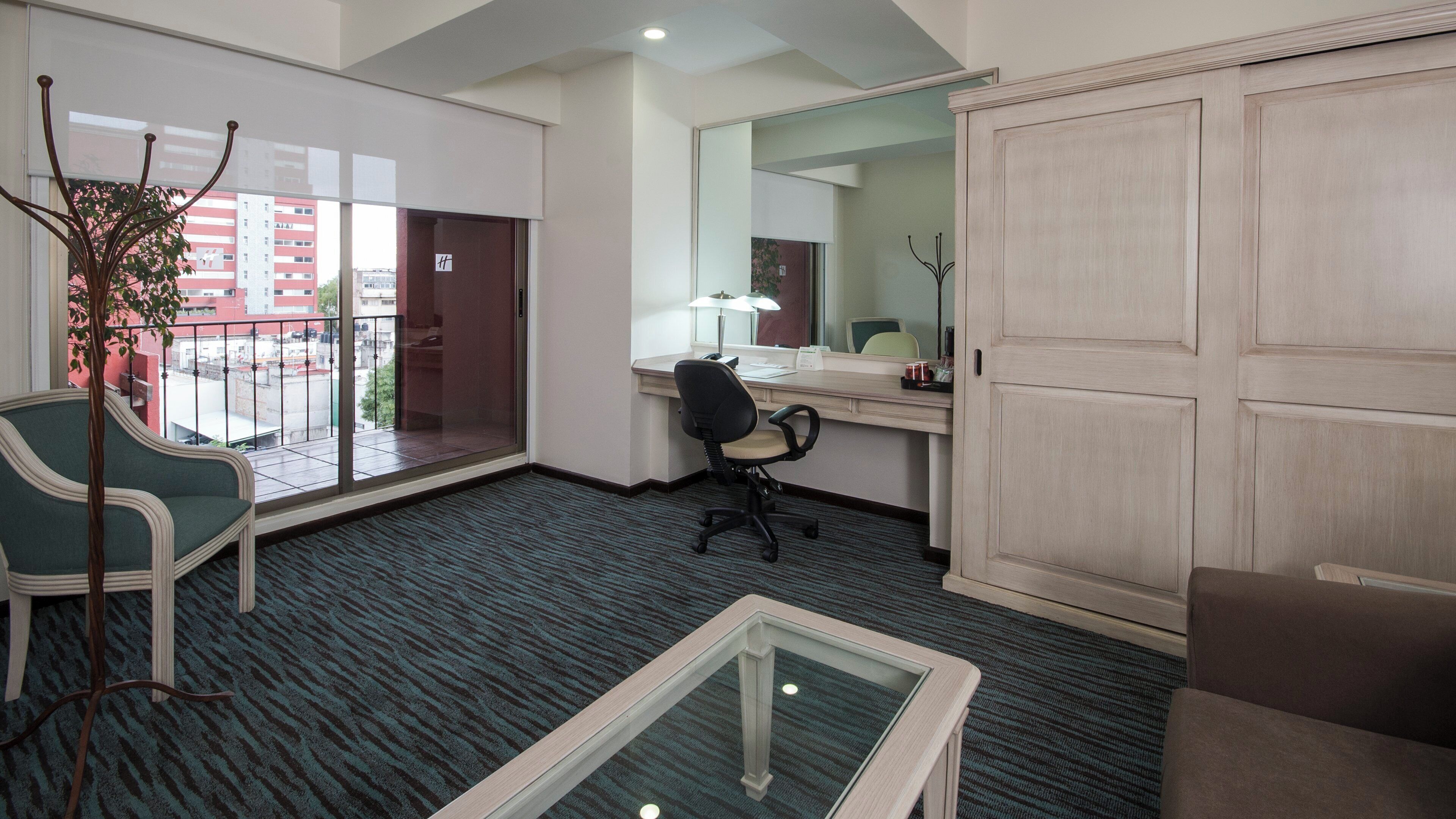 A hotel room with a large sliding door, a desk with a chair, a small table, and a potted plant.
