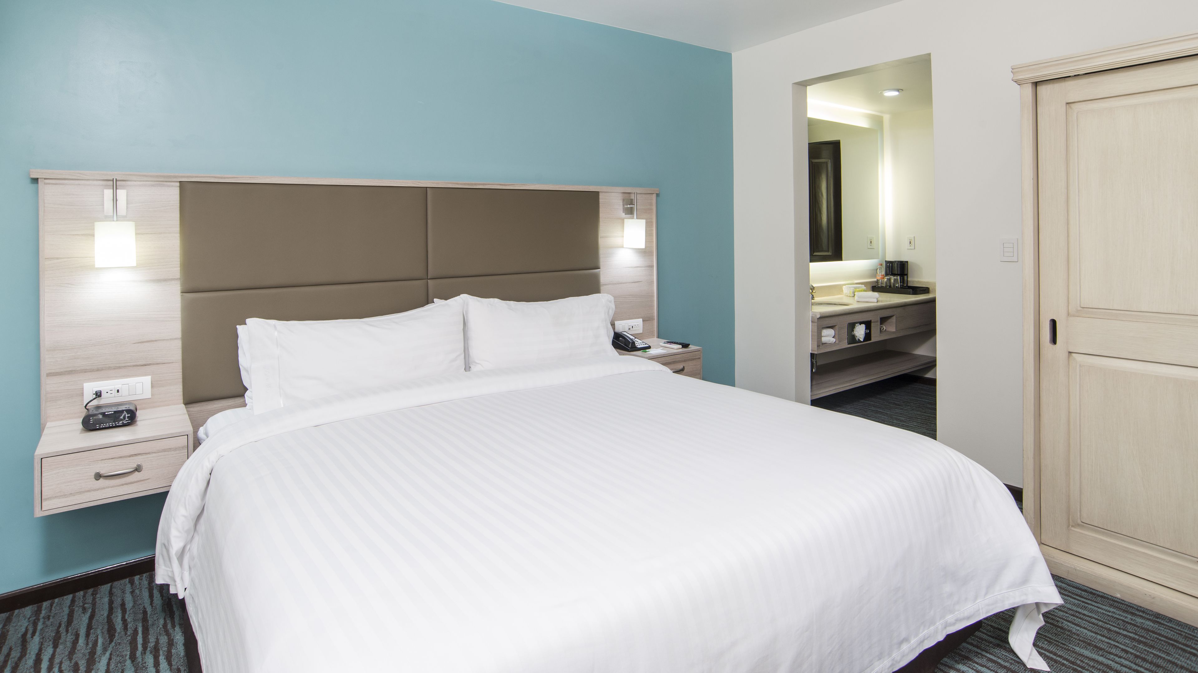 A hotel room with a large bed, a nightstand with a phone and a radio, a vanity with a mirror, and a door leading to another room.