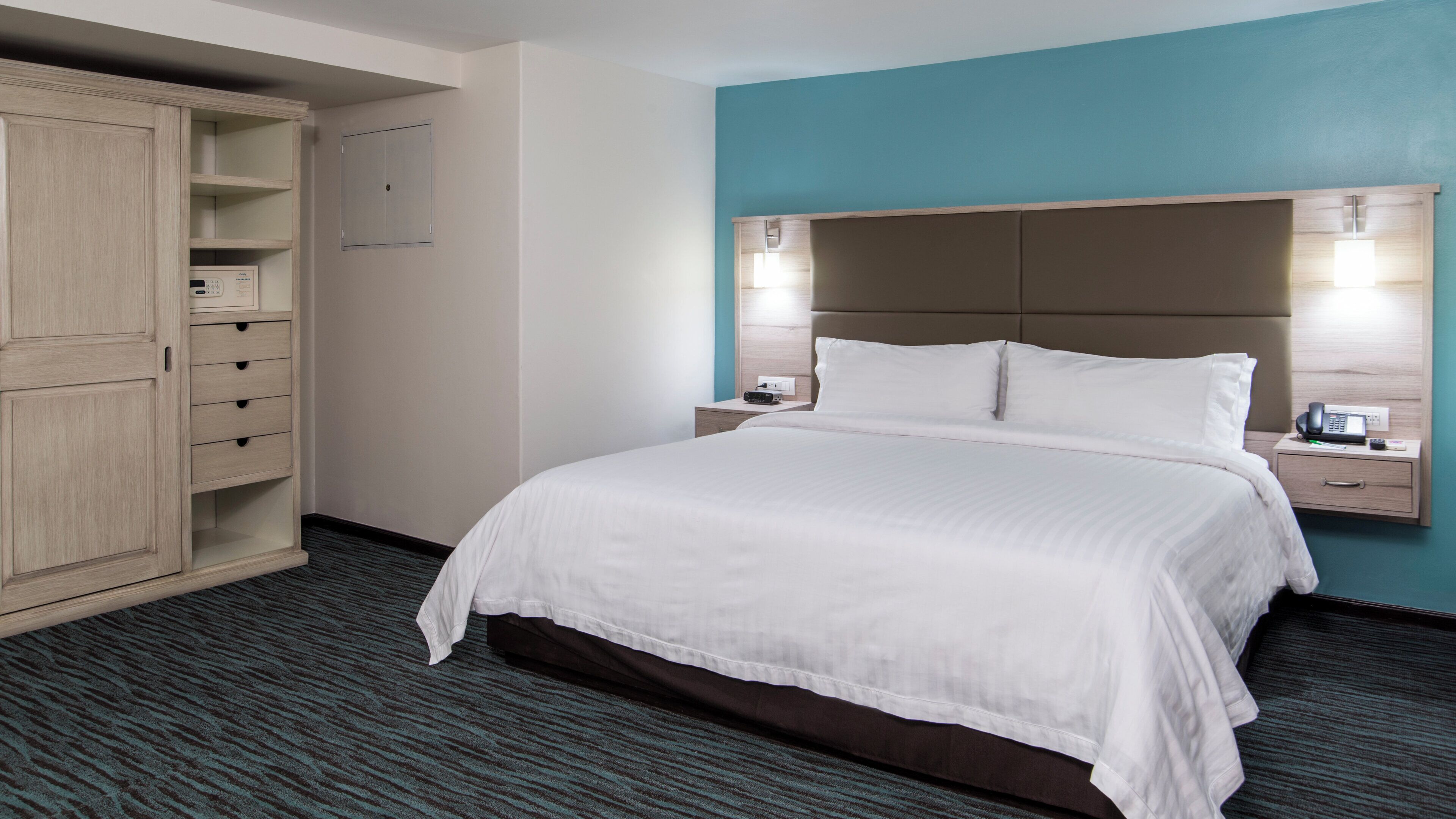 A hotel room with a bed, a wardrobe, a nightstand with a phone, and a wall-mounted light.