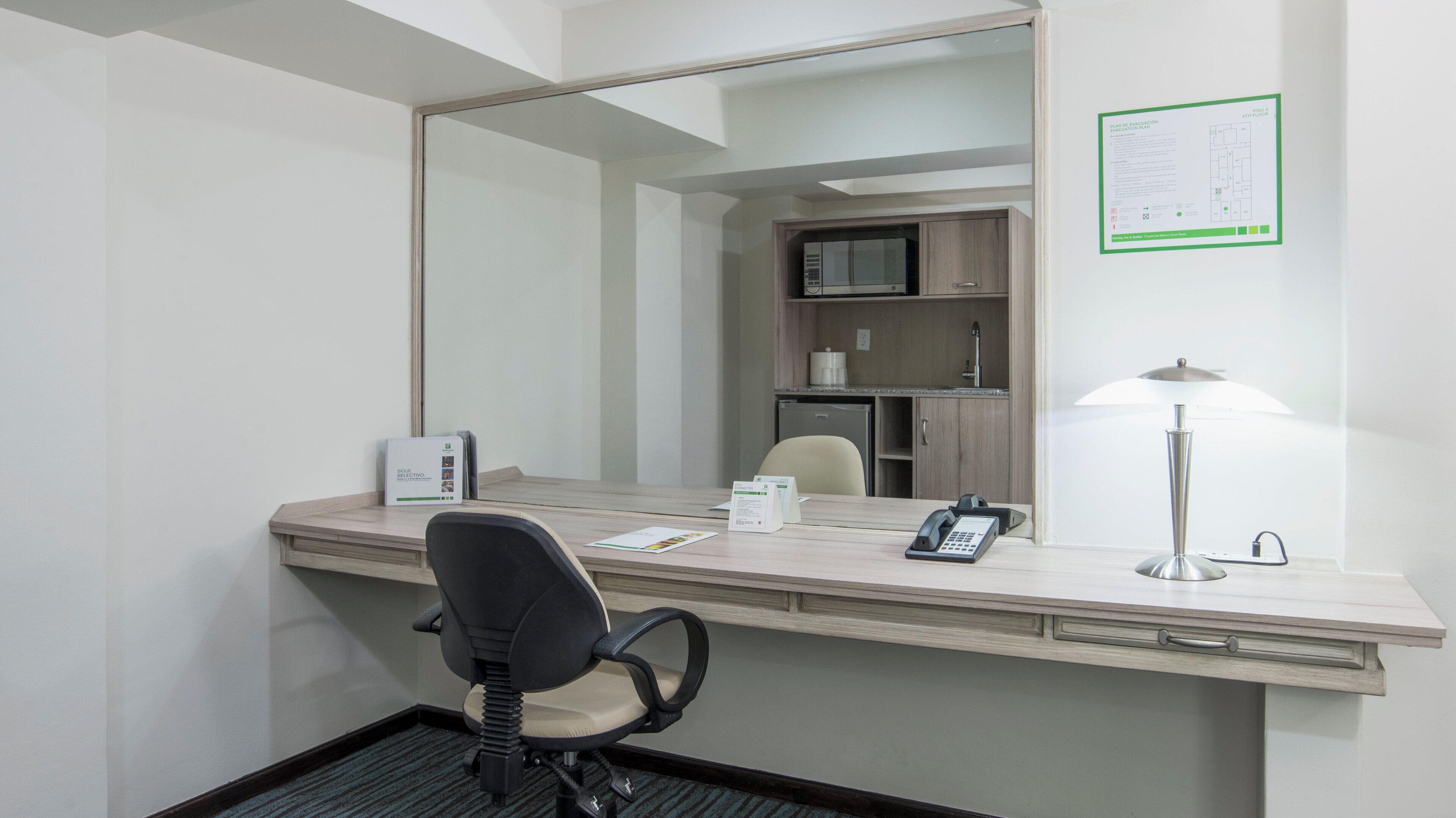 A hotel room with a desk, chair, telephone, lamp, and a mirror.