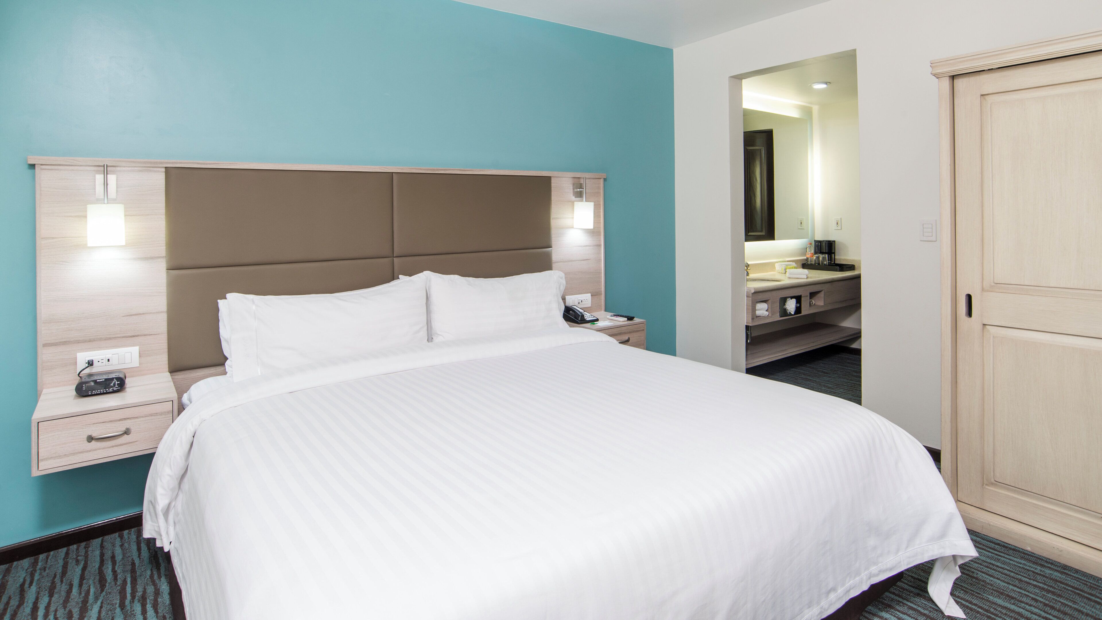A hotel room with a large bed, bedside tables, a nightstand, a bathroom with a sink and mirror, and a wardrobe.