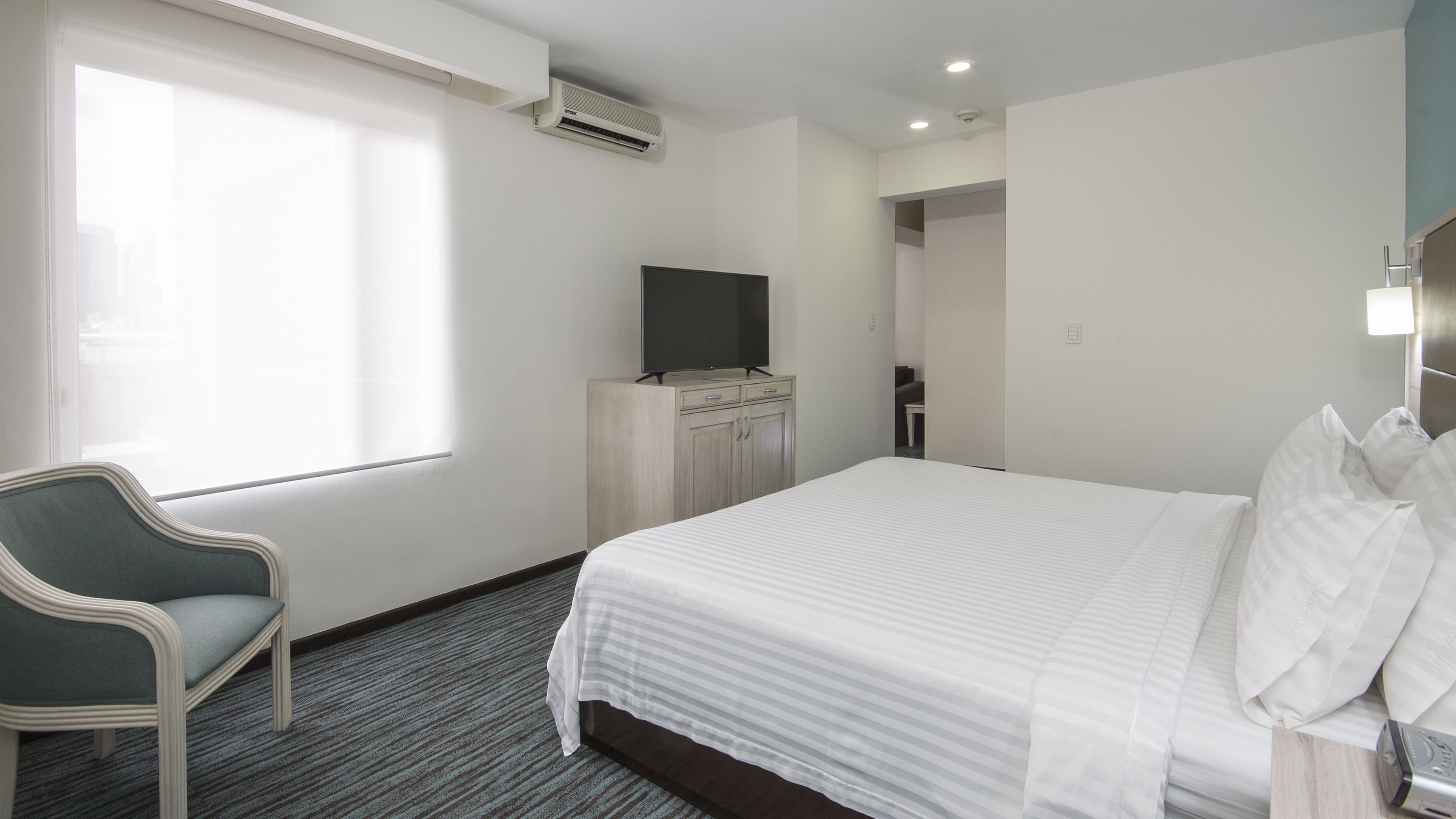 Una habitación de hotel con una cama, un televisor en un mueble, una silla y un ventanal.