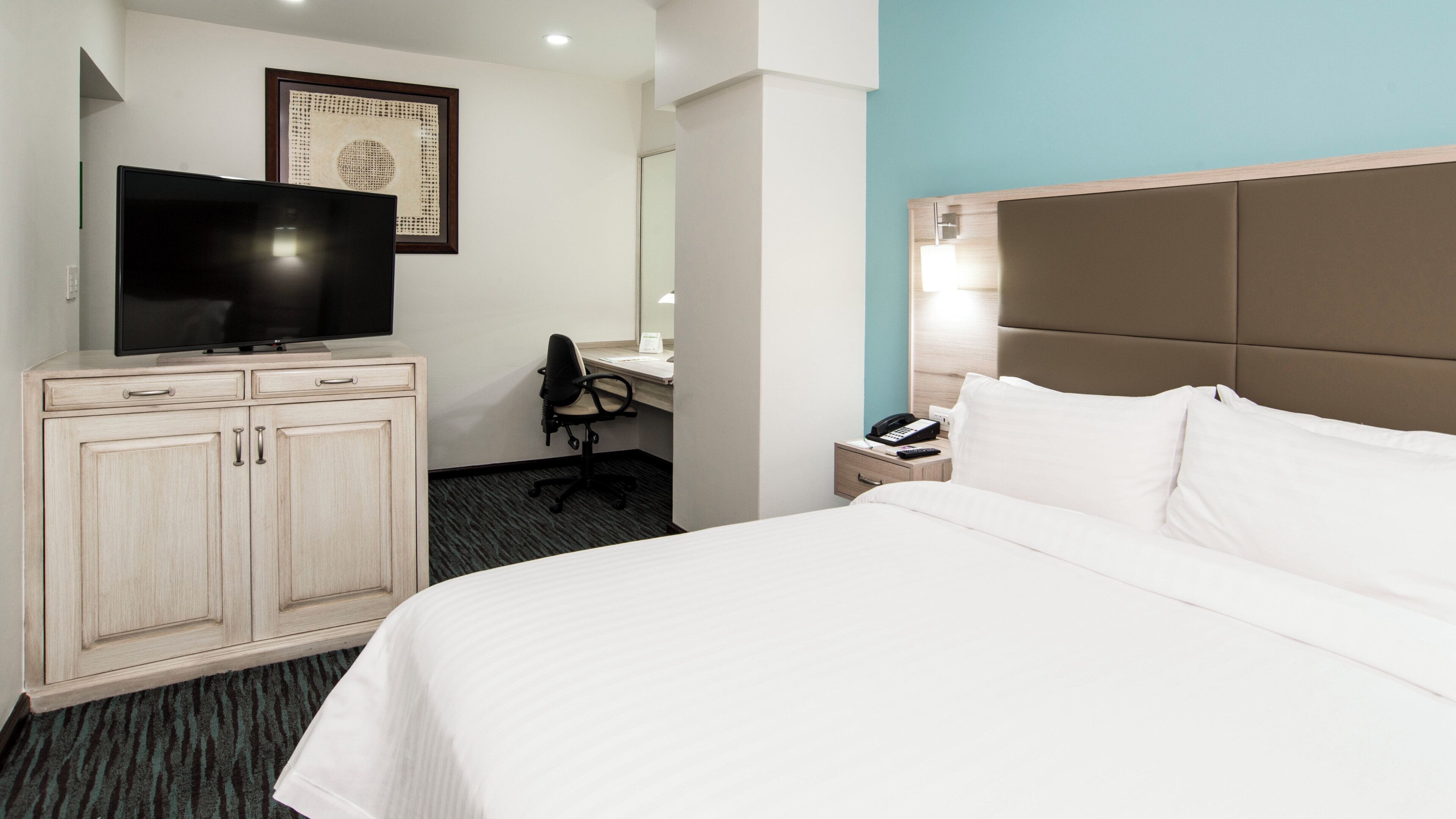 A hotel room with a bed, a desk with a chair, a television, a bedside table with a phone, and a wall-mounted artwork.