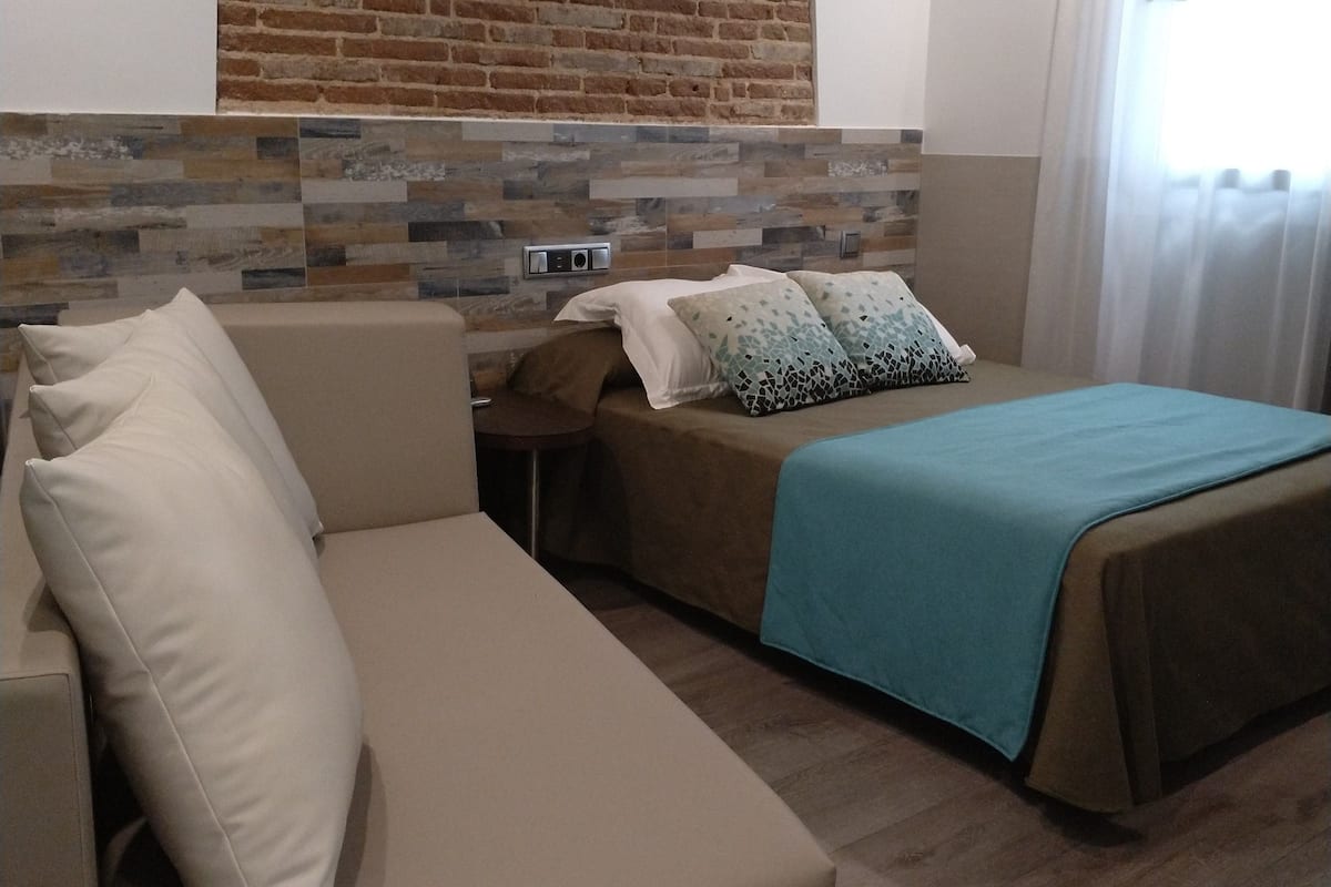 Ein Schlafzimmer mit Backsteinwand, einem Holzkopfteil, einem Bett mit blauer Bettdecke, einem beigen Sofa und einem Nachttisch.