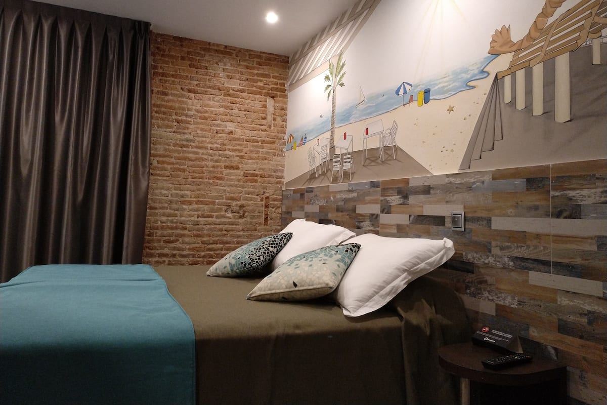 Ein Hotelzimmer mit Backsteinwand, einem Wandgemälde einer Strandansicht, einem Bett mit türkisblau-grüner Bettdecke und einem kleinen Tisch mit Fernbedienung.