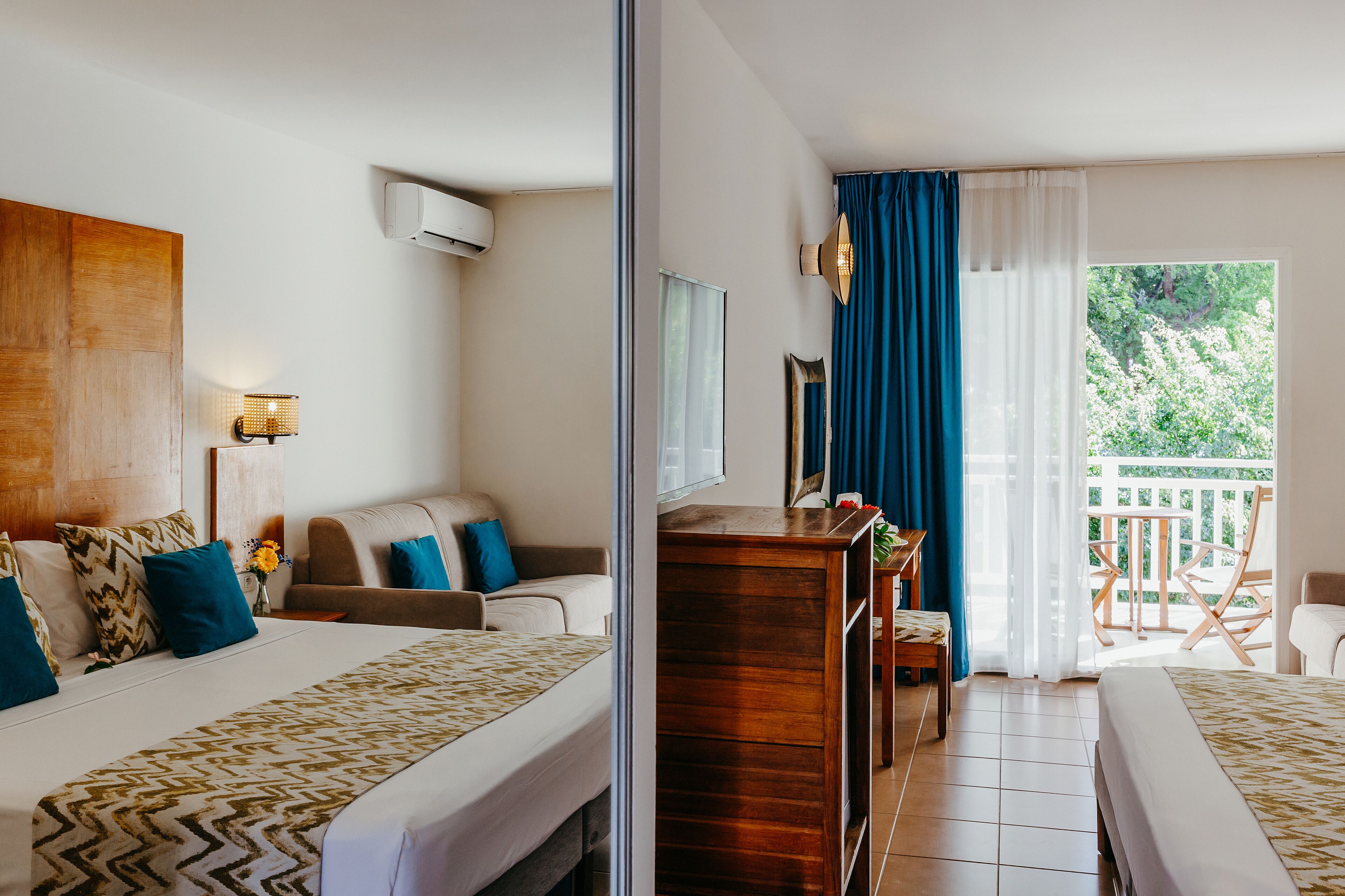 A hotel room with two beds, a wooden dresser, a desk, and a balcony with a view of greenery.