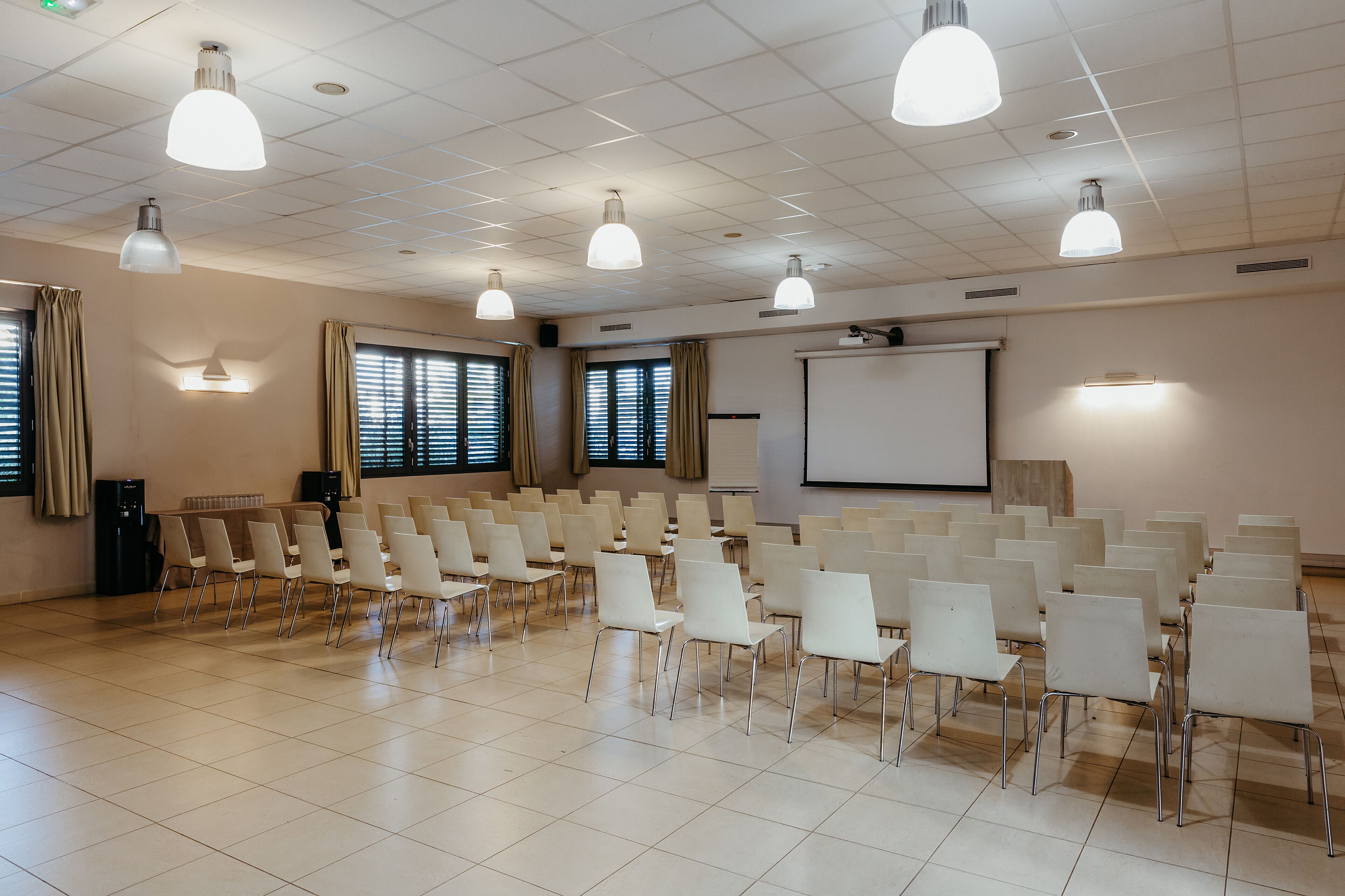 Een vergaderruimte met rijen witte stoelen, een projectiescherm en plafondlampen.
