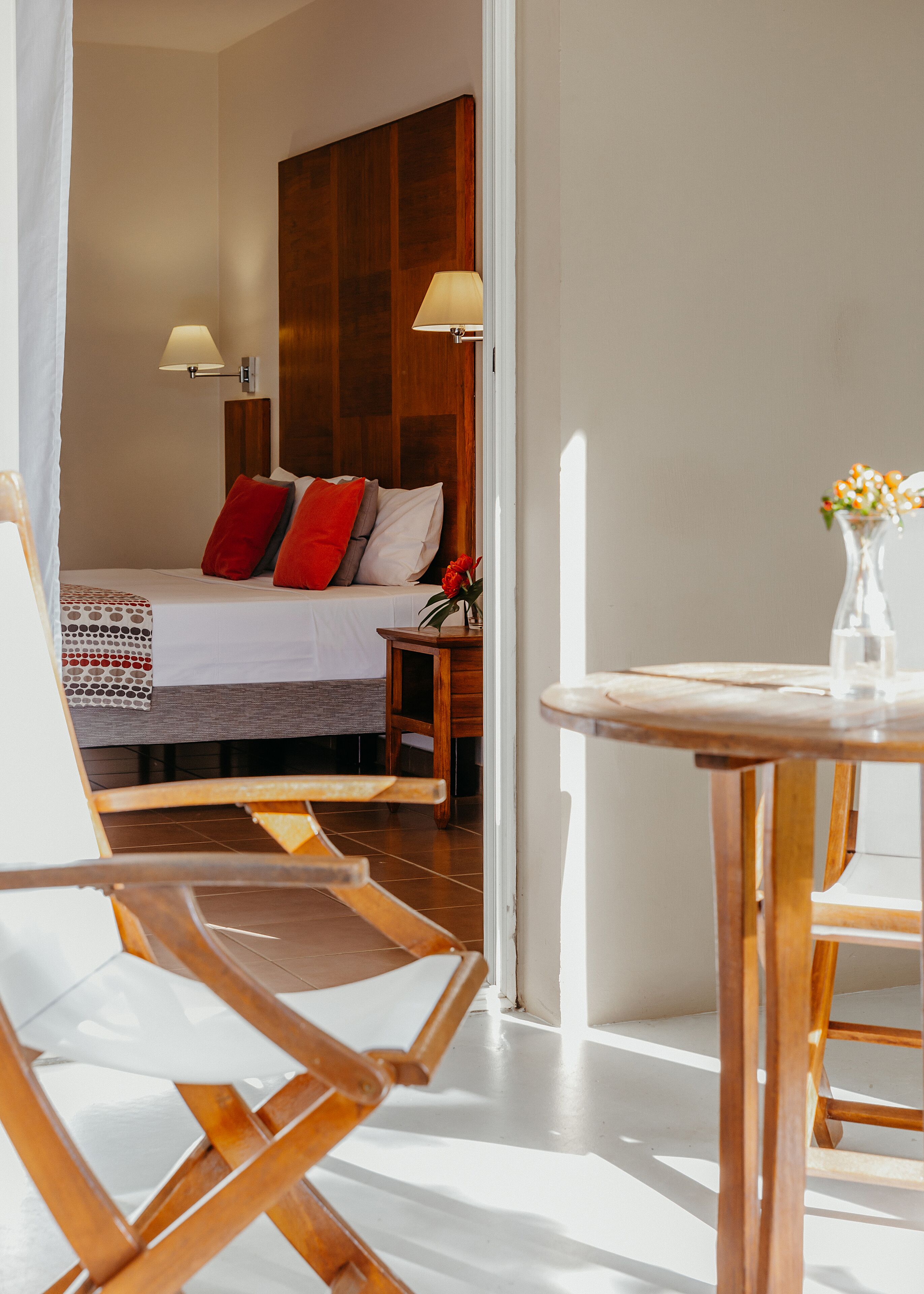 A hotel room with a bed, a wooden headboard, a nightstand, a table with a vase of flowers, and a chair.