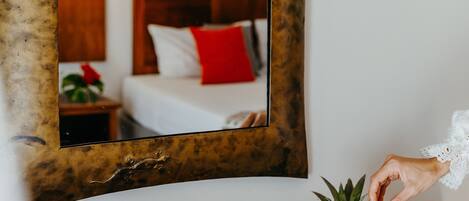 A wooden table with a plate of fruit and a hand reaching for a slice. A mirror reflects a room with a bed and a nightstand.