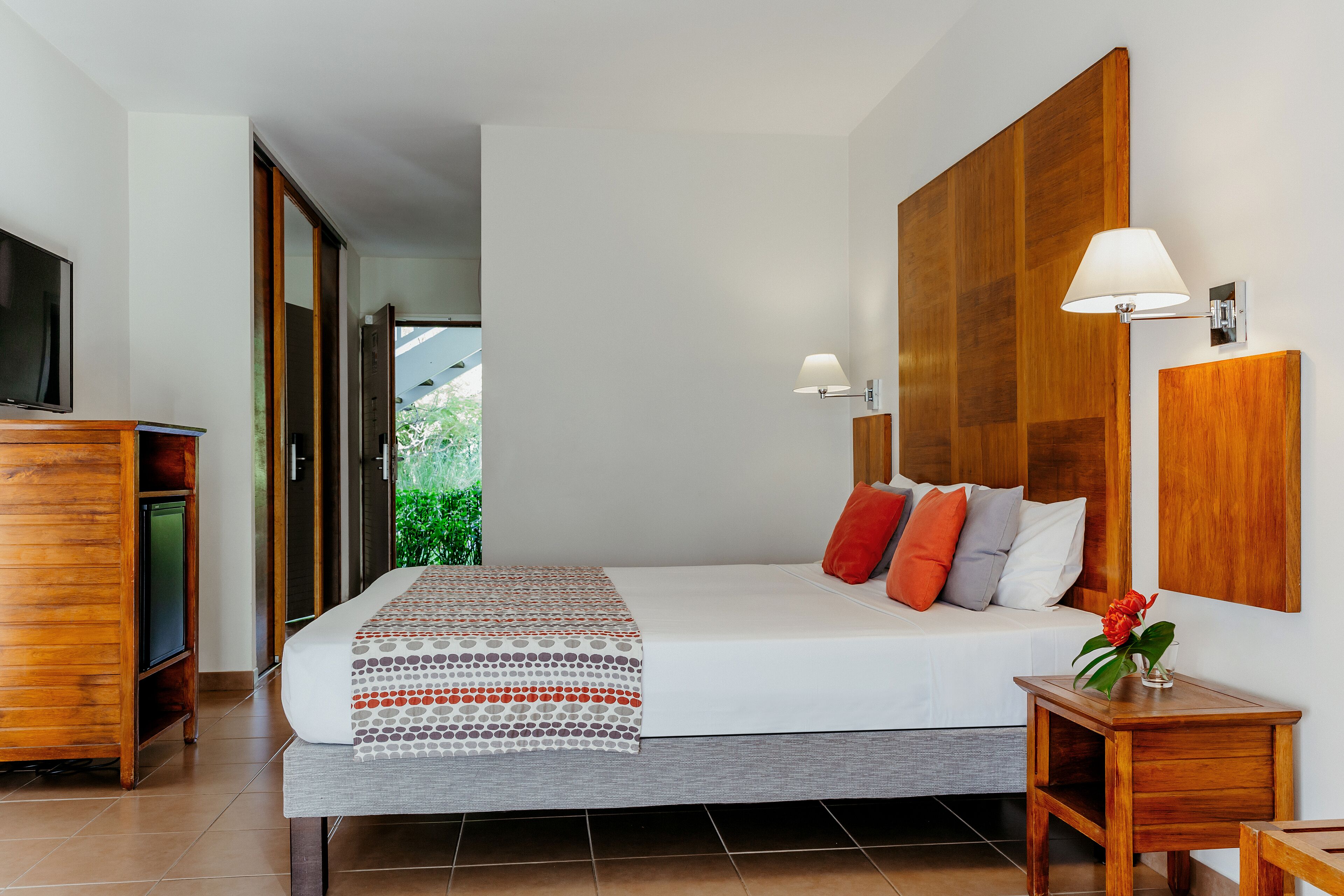 Una habitación de hotel moderna con una cama grande, un cabecero de madera, una lámpara de noche y un televisor sobre un mueble de madera.